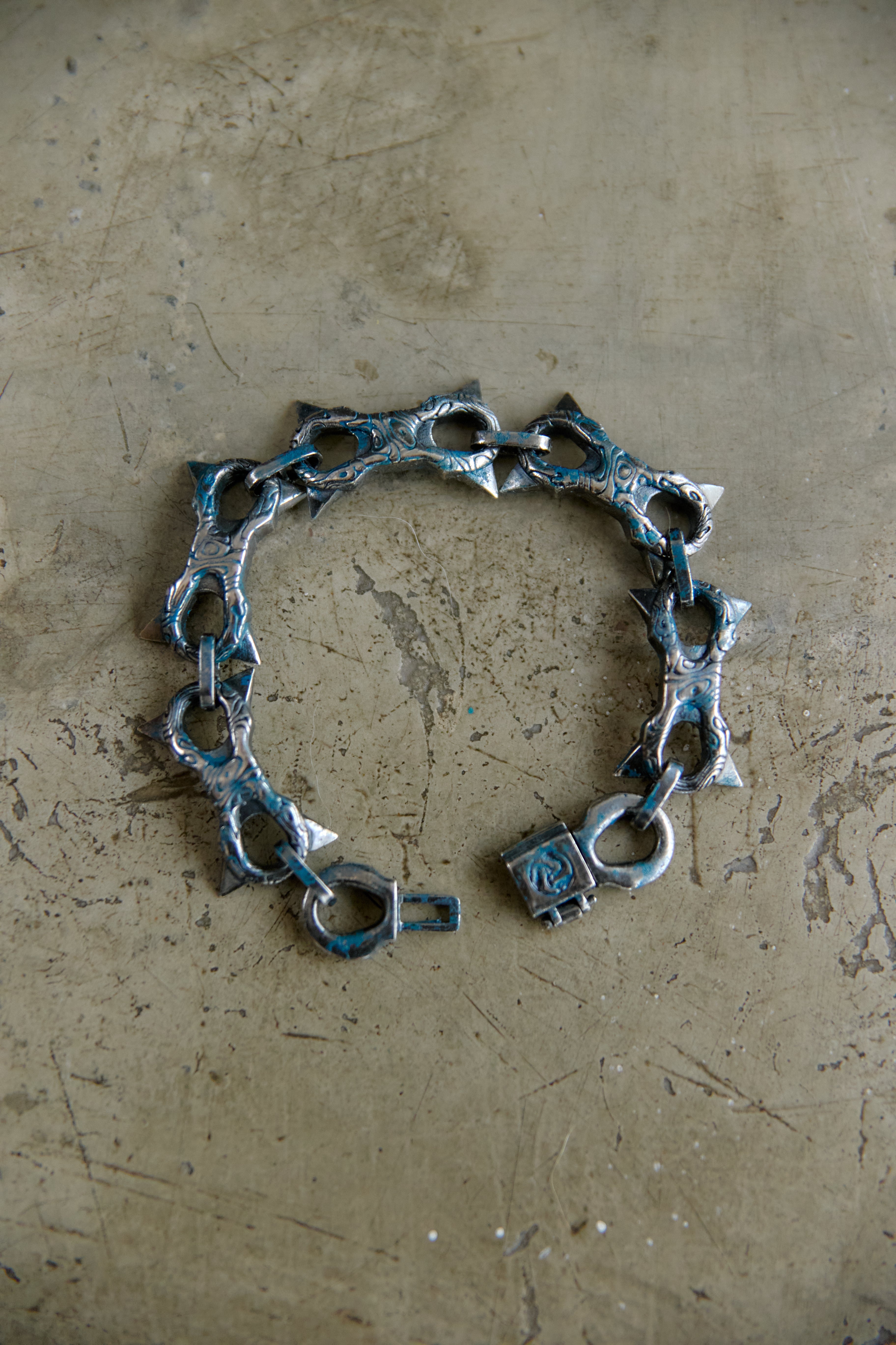 A handmade silver-toned chain bracelet with triangular cutouts and a padlock clasp, with blueish patina oxidation on its surface, displayed on a textured, scratched beige surface.