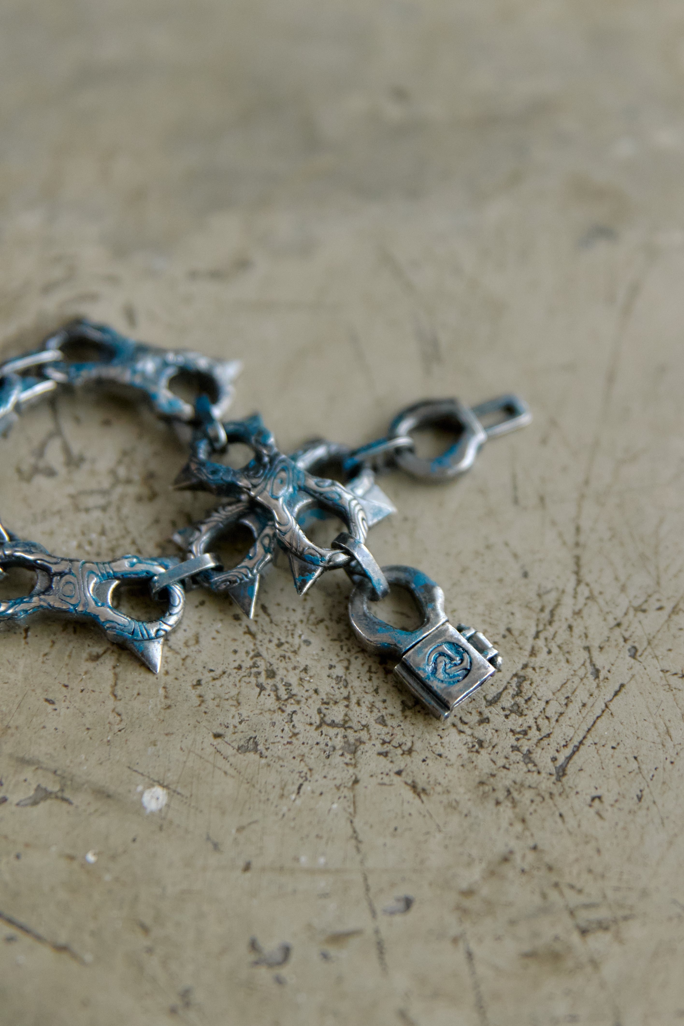 A close-up of a metallic silver chain bracelet with sharp spikes and a padlock charm, resting on a textured, scratched beige surface.