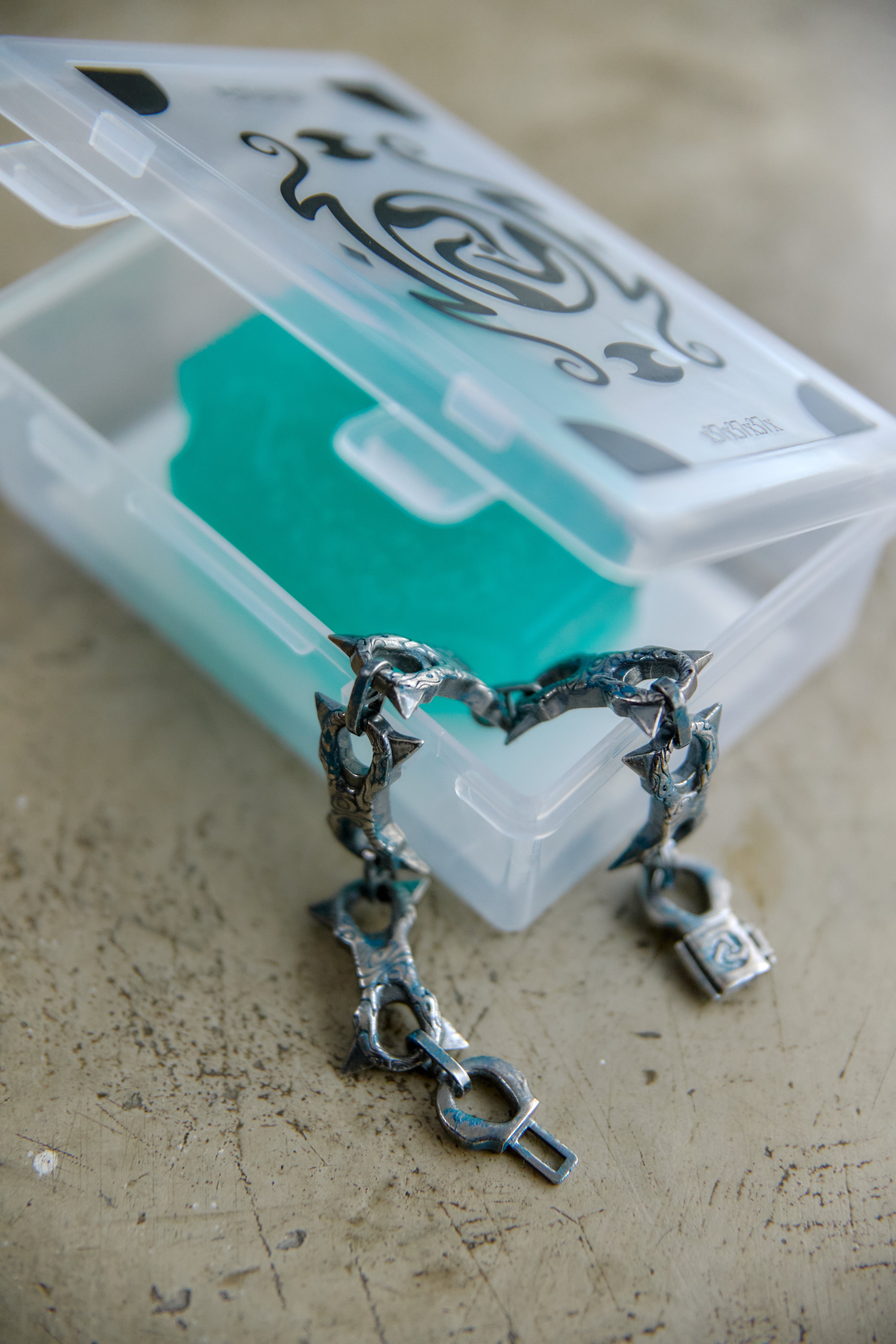 A textured silver-tone metal bracelet with a heart-shaped link design and a small padlock charm, displayed in front of a clear plastic storage case containing a turquoise soap block on a beige surface.