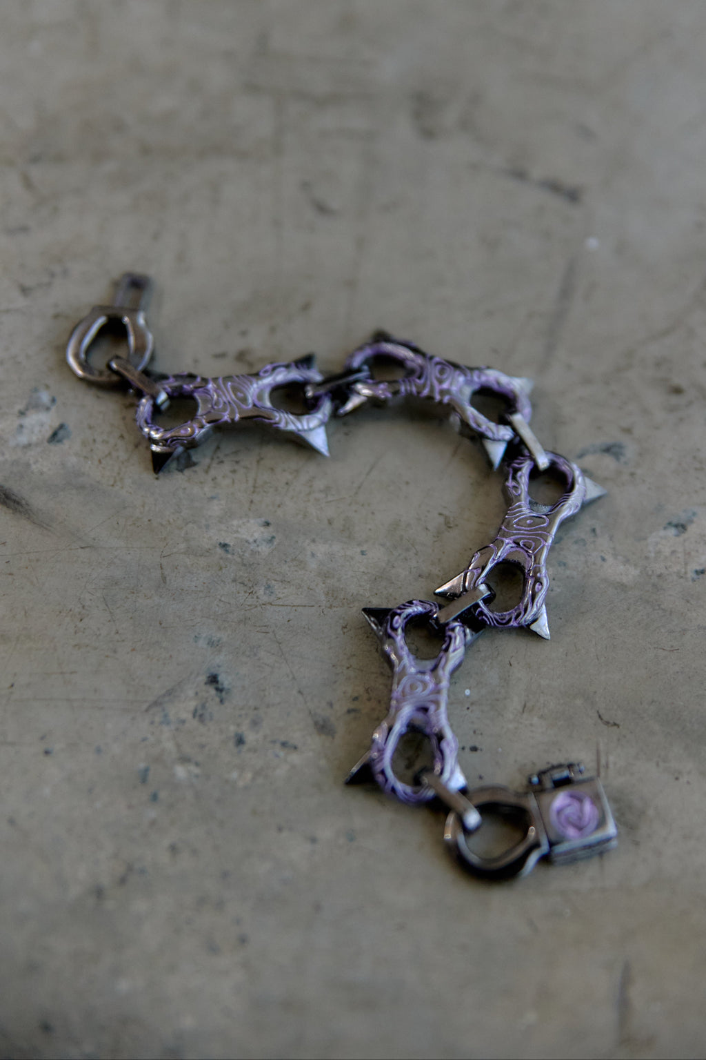 A gothic-style spiked chain bracelet with a dark metallic finish and a small pink heart charm, resting on a textured concrete surface.