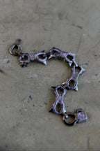 A gothic-style spiked chain bracelet with a dark metallic finish and a small pink heart charm, resting on a textured concrete surface.