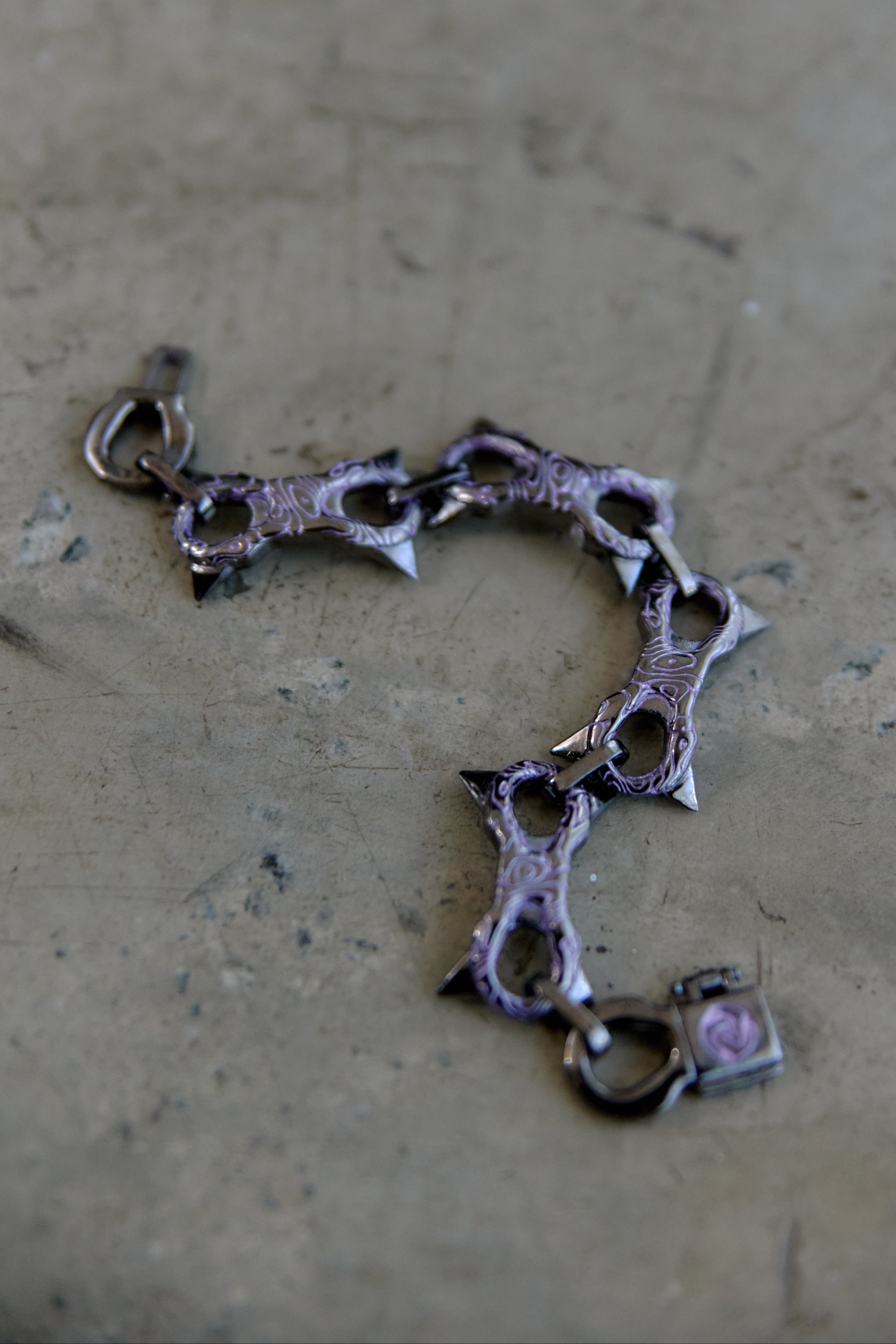 A gothic-style spiked chain bracelet with a dark metallic finish and a small pink heart charm, resting on a textured concrete surface.