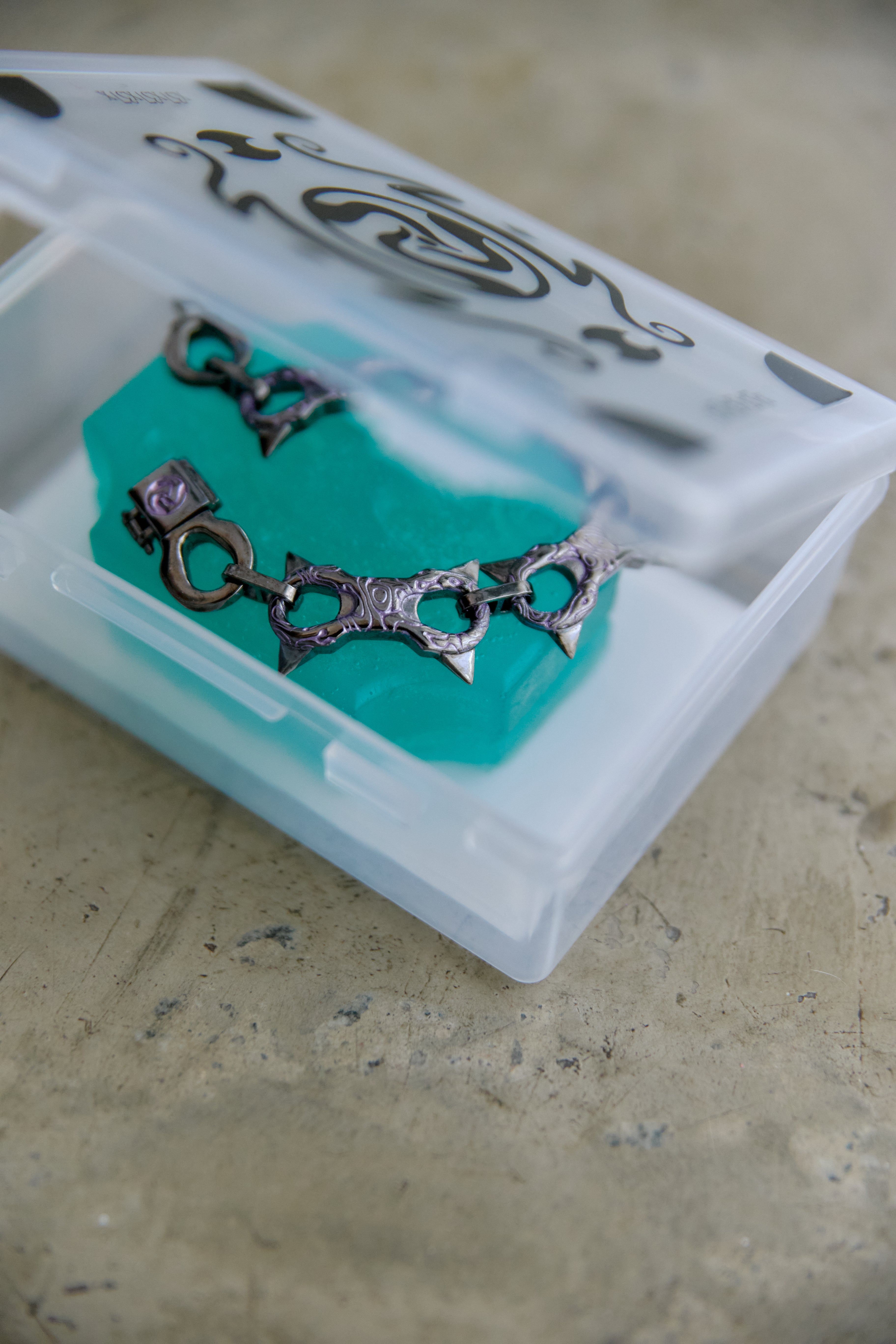 A silver-toned chain link bracelet with geometric and tribal-inspired cutouts, resting on a bright turquoise block inside a clear plastic storage case with a patterned lid, set on a textured beige surface.
