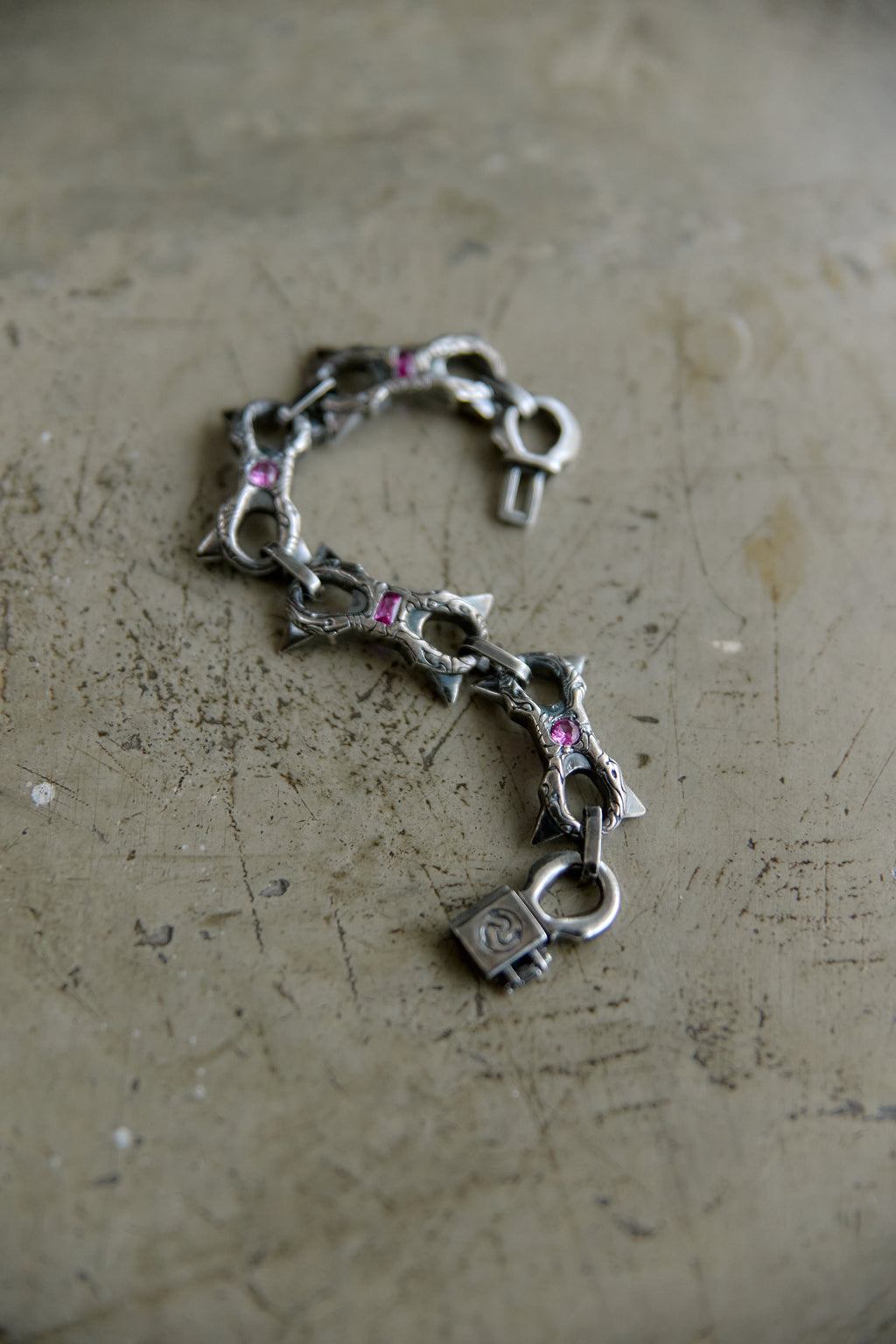 A silver-toned chain bracelet featuring stylized links shaped like fish and a padlock charm, accented with small pink gemstones, displayed on a textured concrete surface.