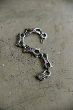 A silver-toned chain bracelet featuring stylized links shaped like fish and a padlock charm, accented with small pink gemstones, displayed on a textured concrete surface.