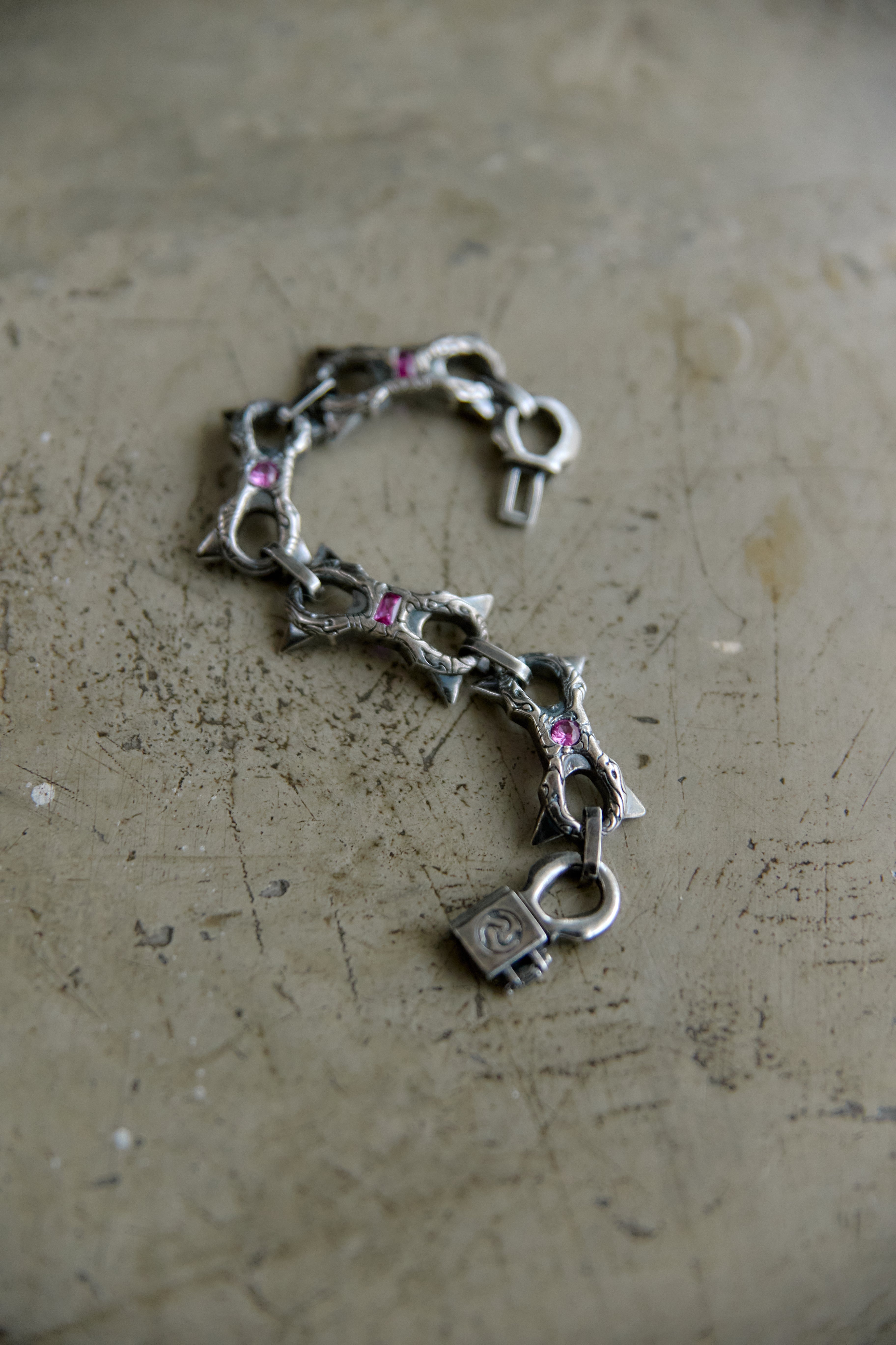 A silver-toned chain bracelet featuring stylized links shaped like fish and a padlock charm, accented with small pink gemstones, displayed on a textured concrete surface.