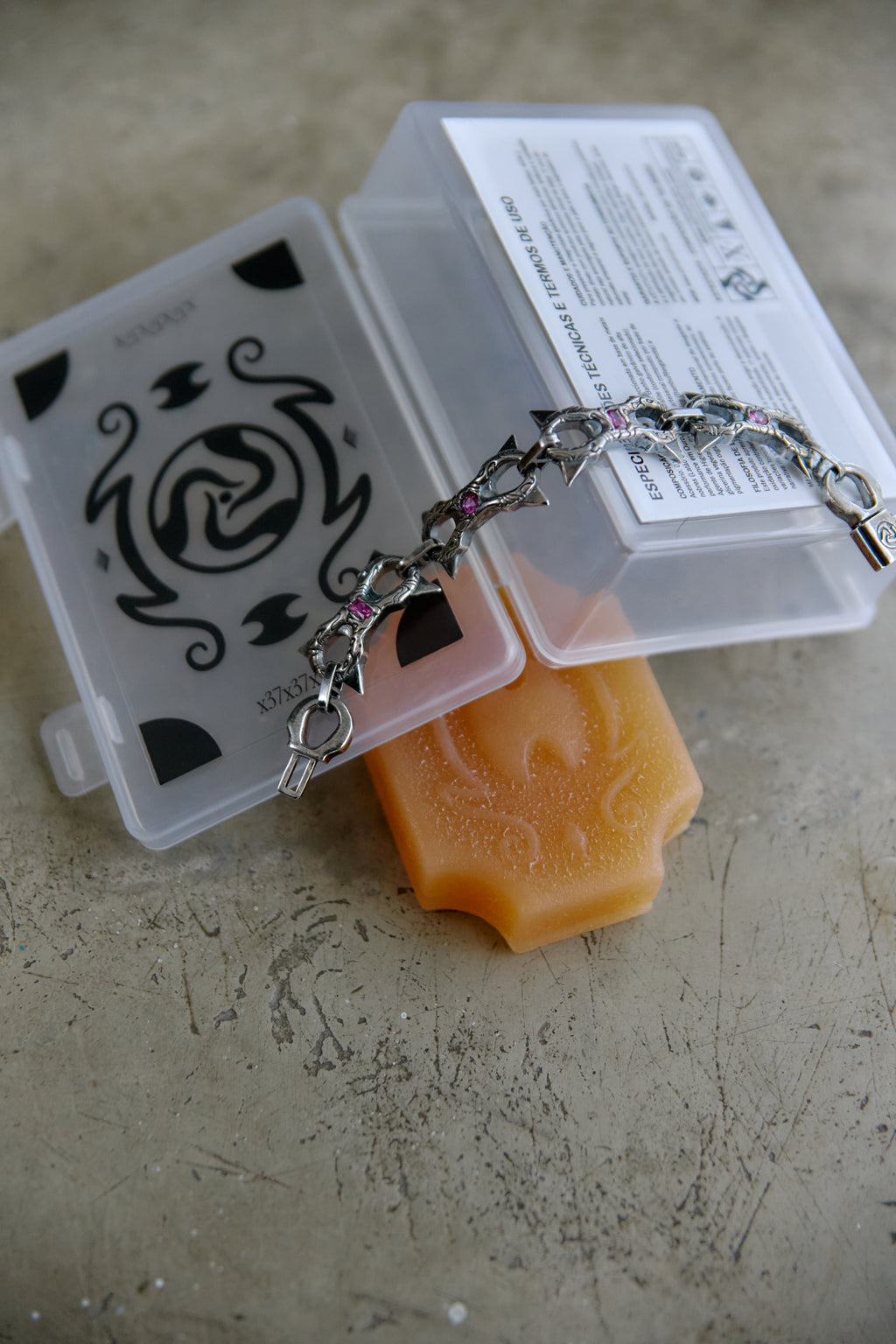 A silver chain bracelet with small purple gemstone accents draped over a translucent orange bar of soap, placed on top of two clear plastic storage boxes with printed labels, on a textured beige surface.