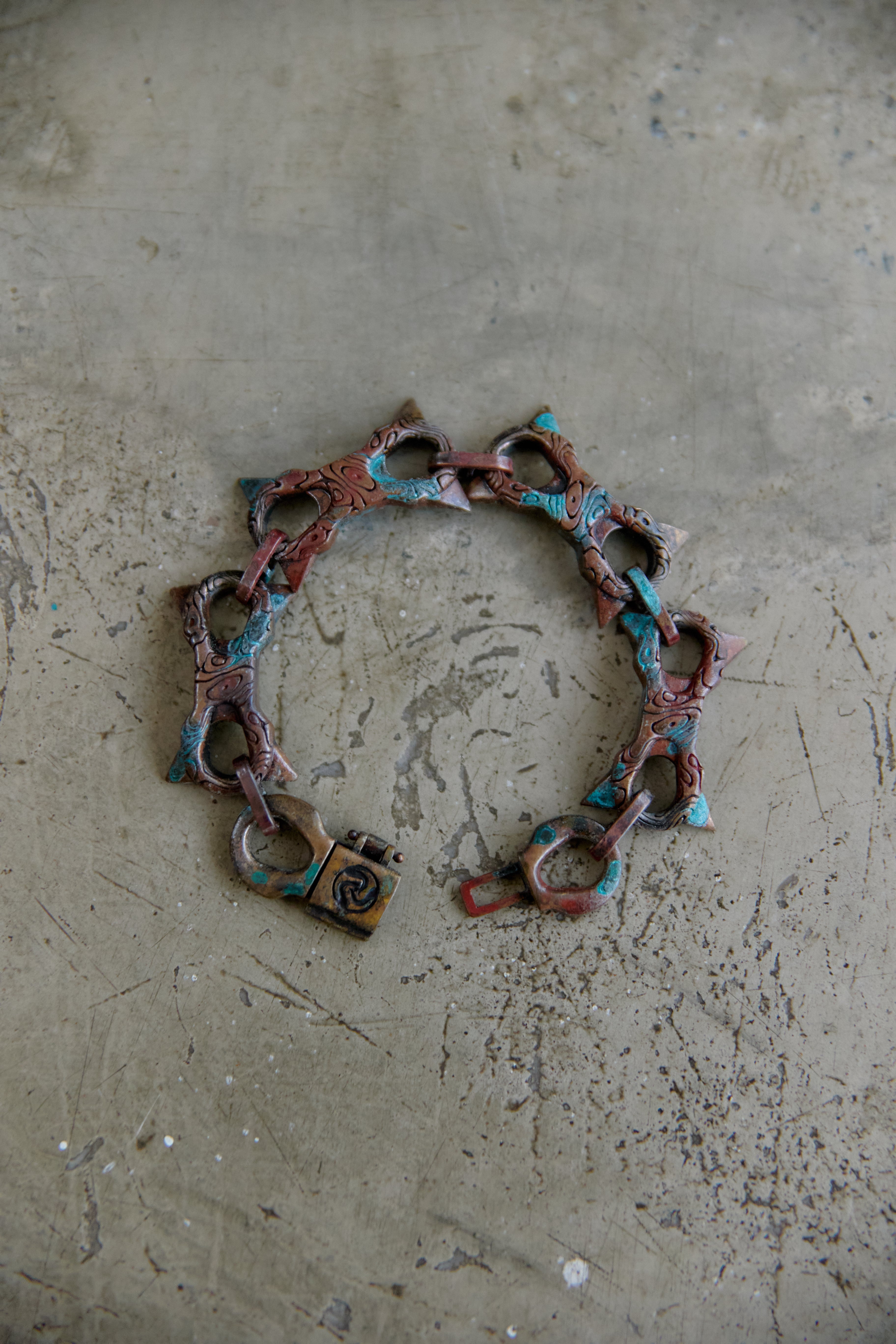 A handmade link bracelet with a padlock clasp, featuring a distressed copper and turquoise patina finish, displayed on a textured concrete surface.