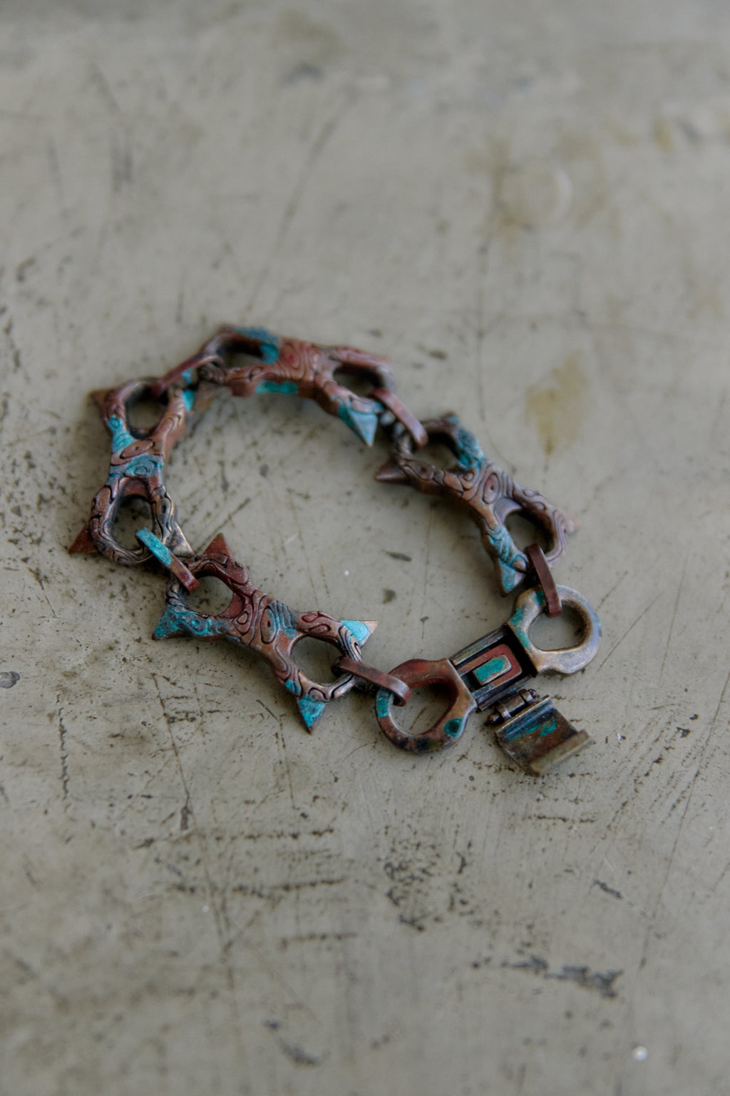 A handmade link bracelet made of oxidized copper-toned metal with turquoise and green inlay details, resting on a textured concrete surface.