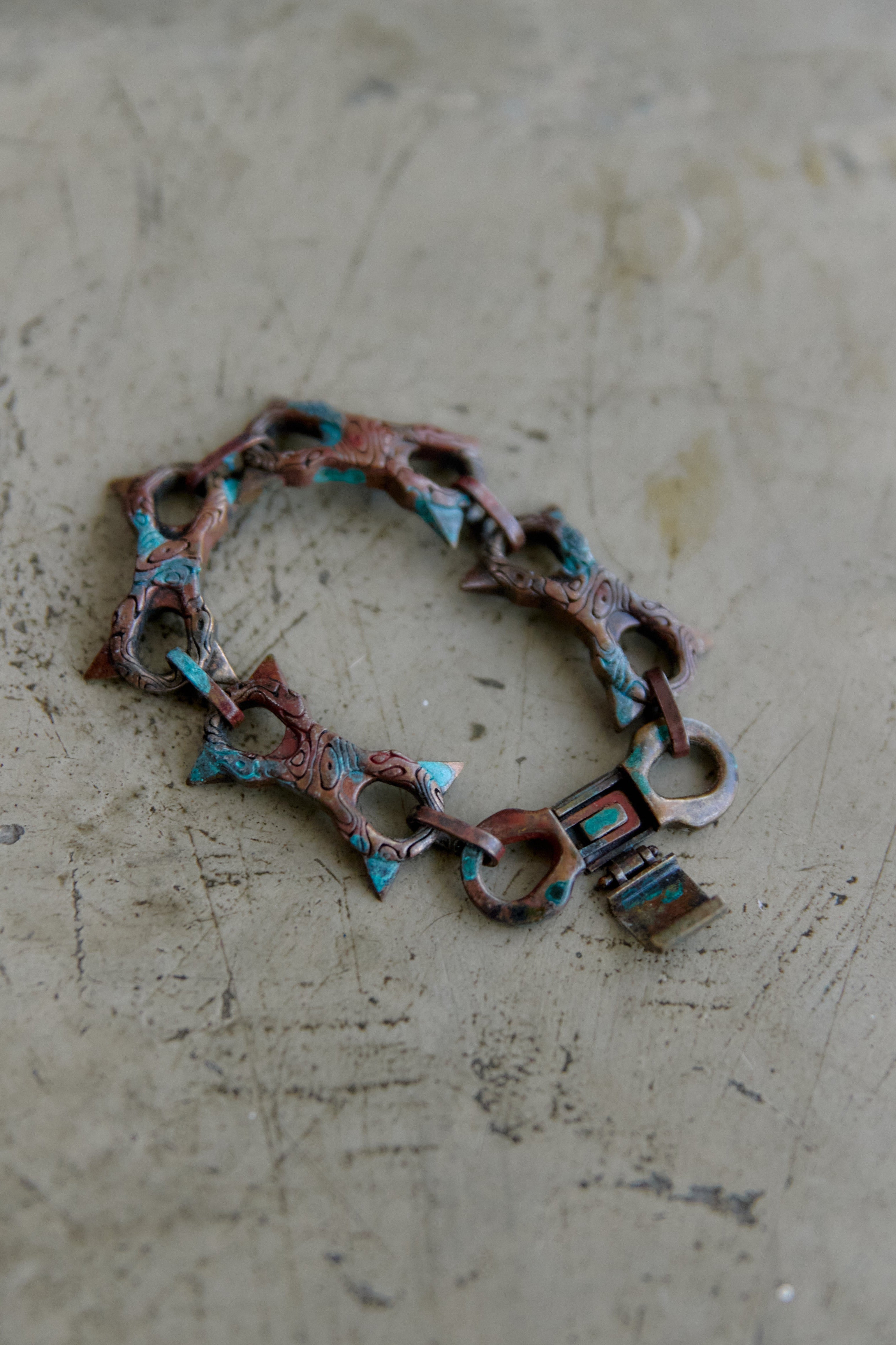 A handmade link bracelet made of oxidized copper-toned metal with turquoise and green inlay details, resting on a textured concrete surface.