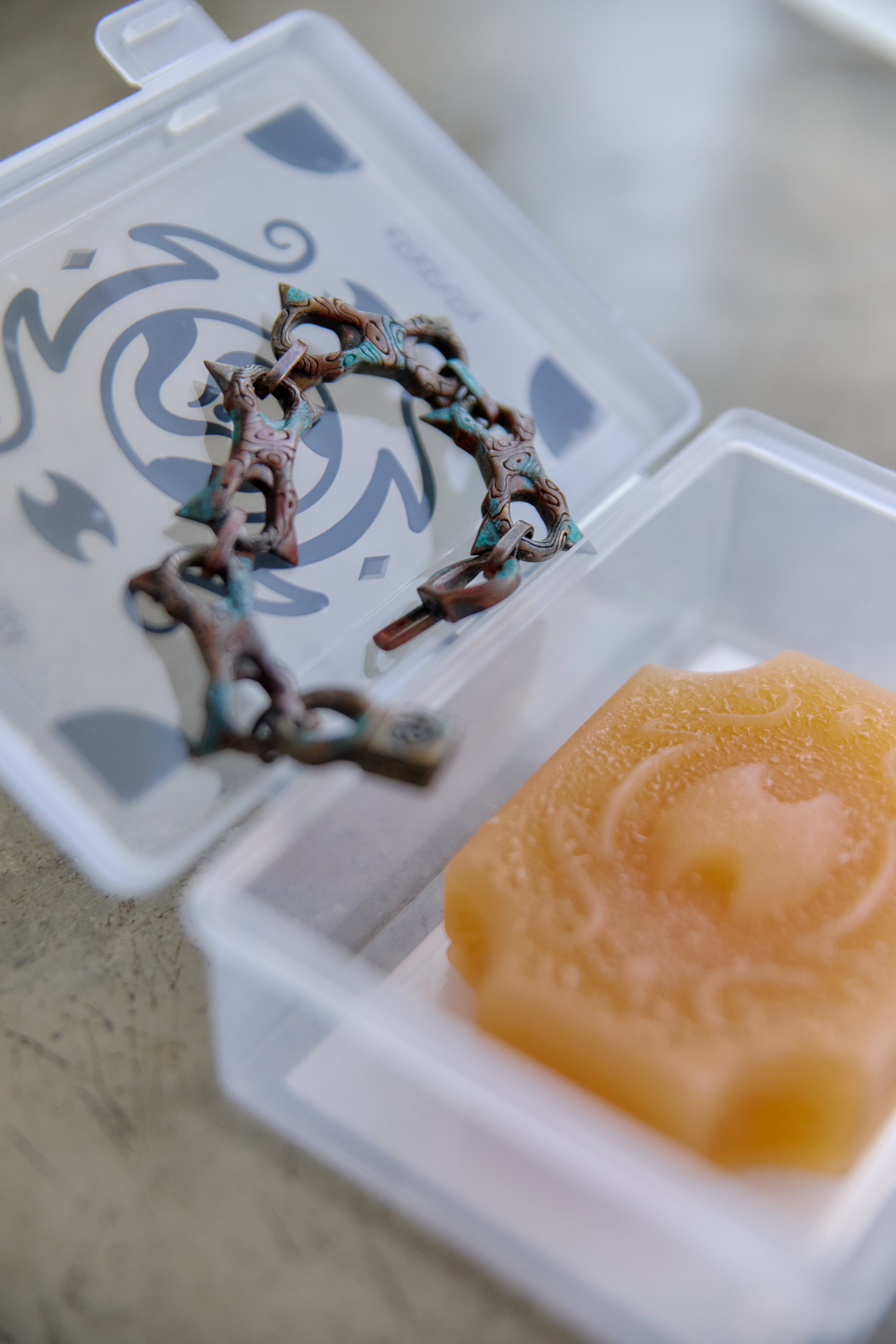 A close-up of a small clear plastic jewelry case opened to show a tarnished bronze chain bracelet inside, next to a translucent amber-colored wax melt or soap piece on a neutral surface.