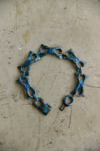 A handmade link bracelet made of textured metal with a distressed blue and bronze patina, resting on a scratched concrete surface.