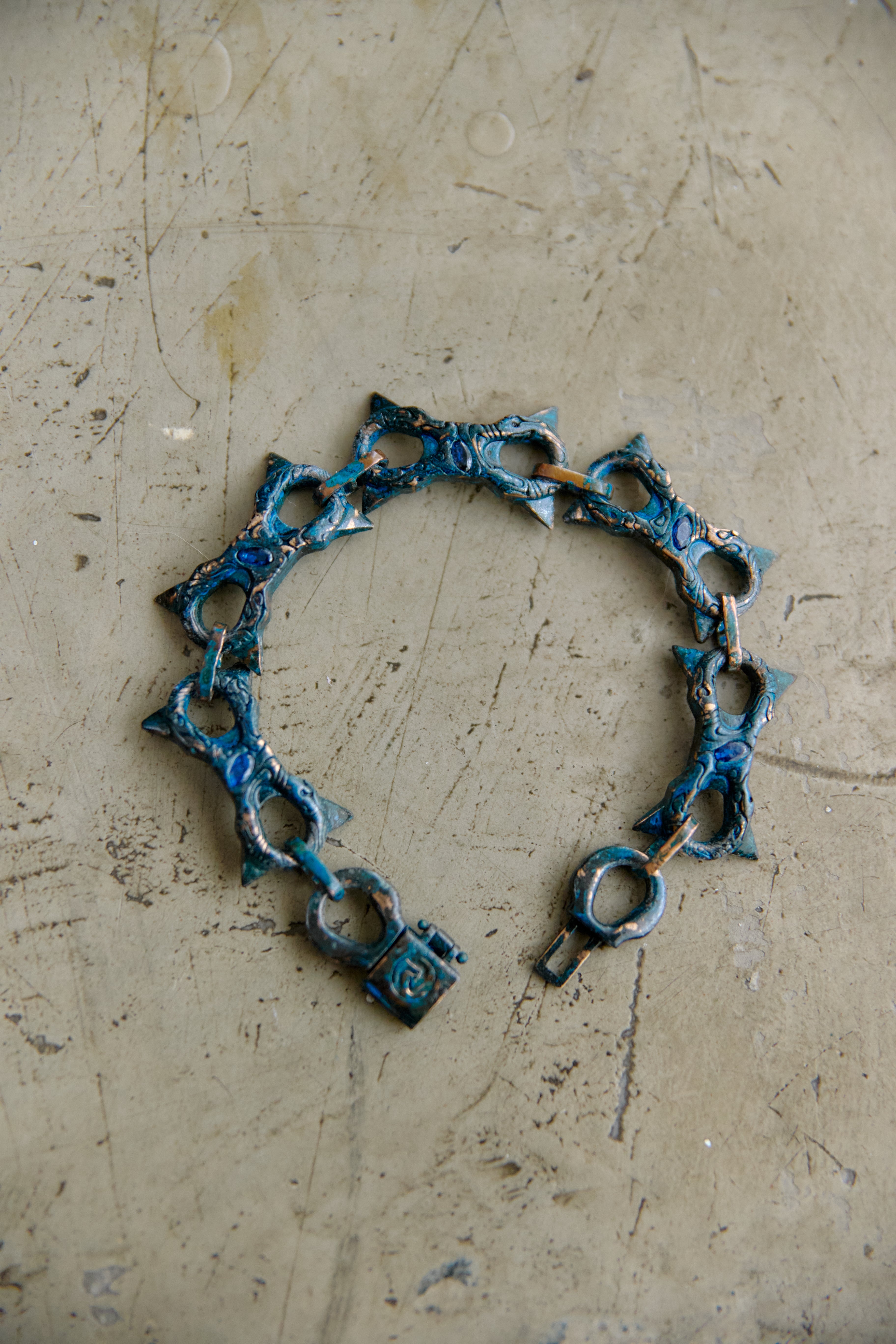 A handmade link bracelet made of textured metal with a distressed blue and bronze patina, resting on a scratched concrete surface.