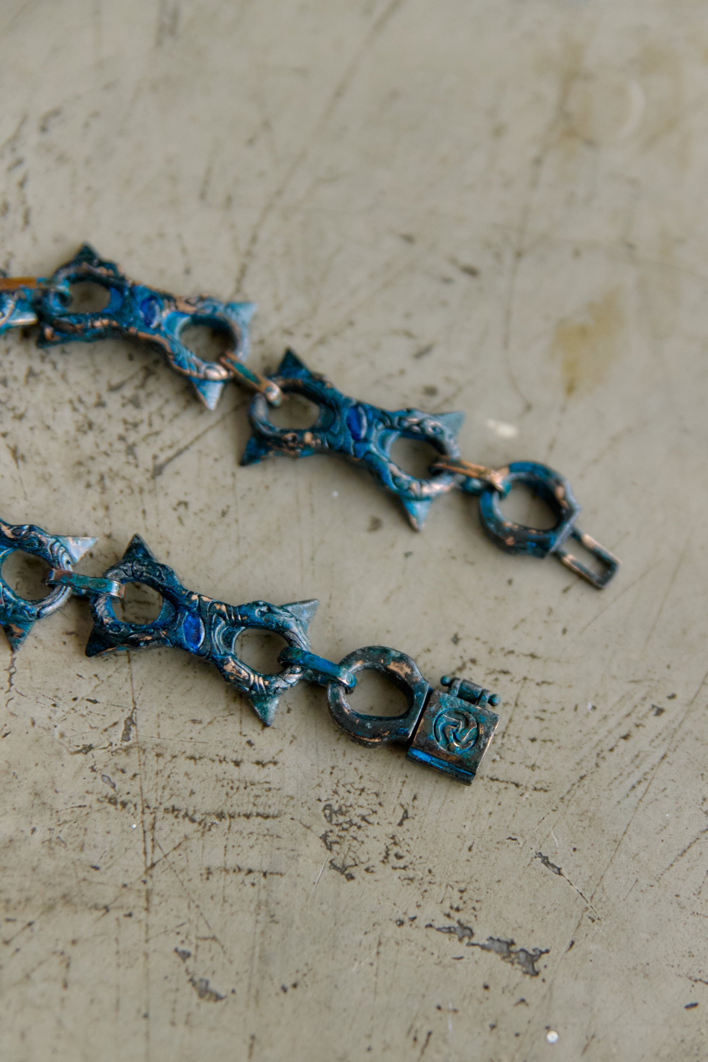 A close-up of a textured metal chain with ornate, openwork links in a distressed blue and silver finish, resting on a scratched, light gray concrete-like surface.