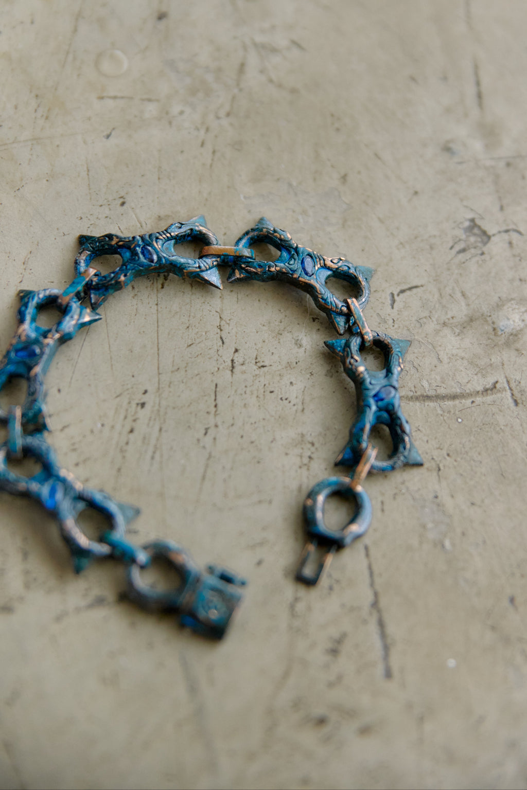 A handmade link bracelet with a rustic, oxidized blue and brown patina, displayed on a textured beige concrete surface.