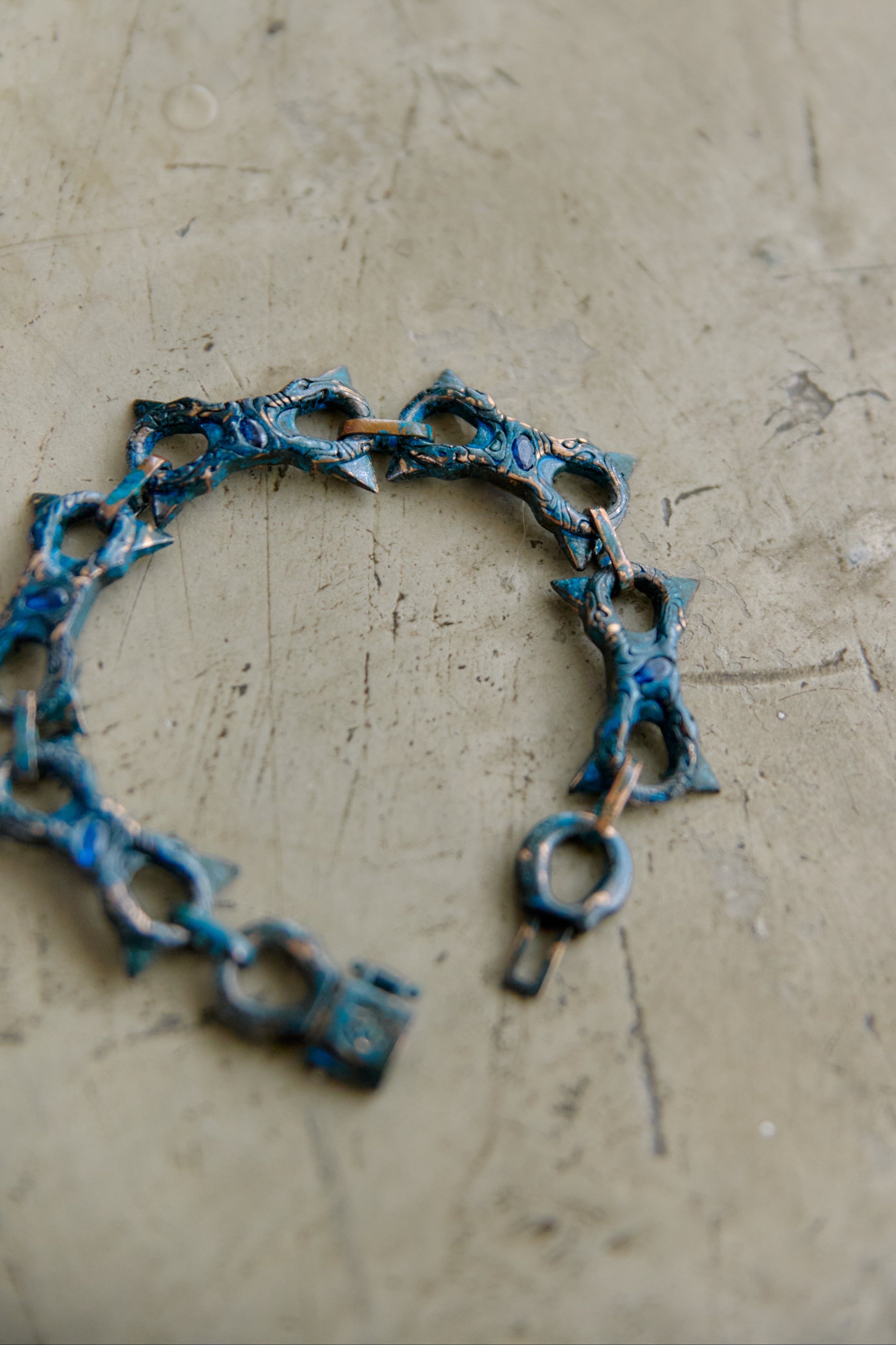 A handmade link bracelet with a rustic, oxidized blue and brown patina, displayed on a textured beige concrete surface.