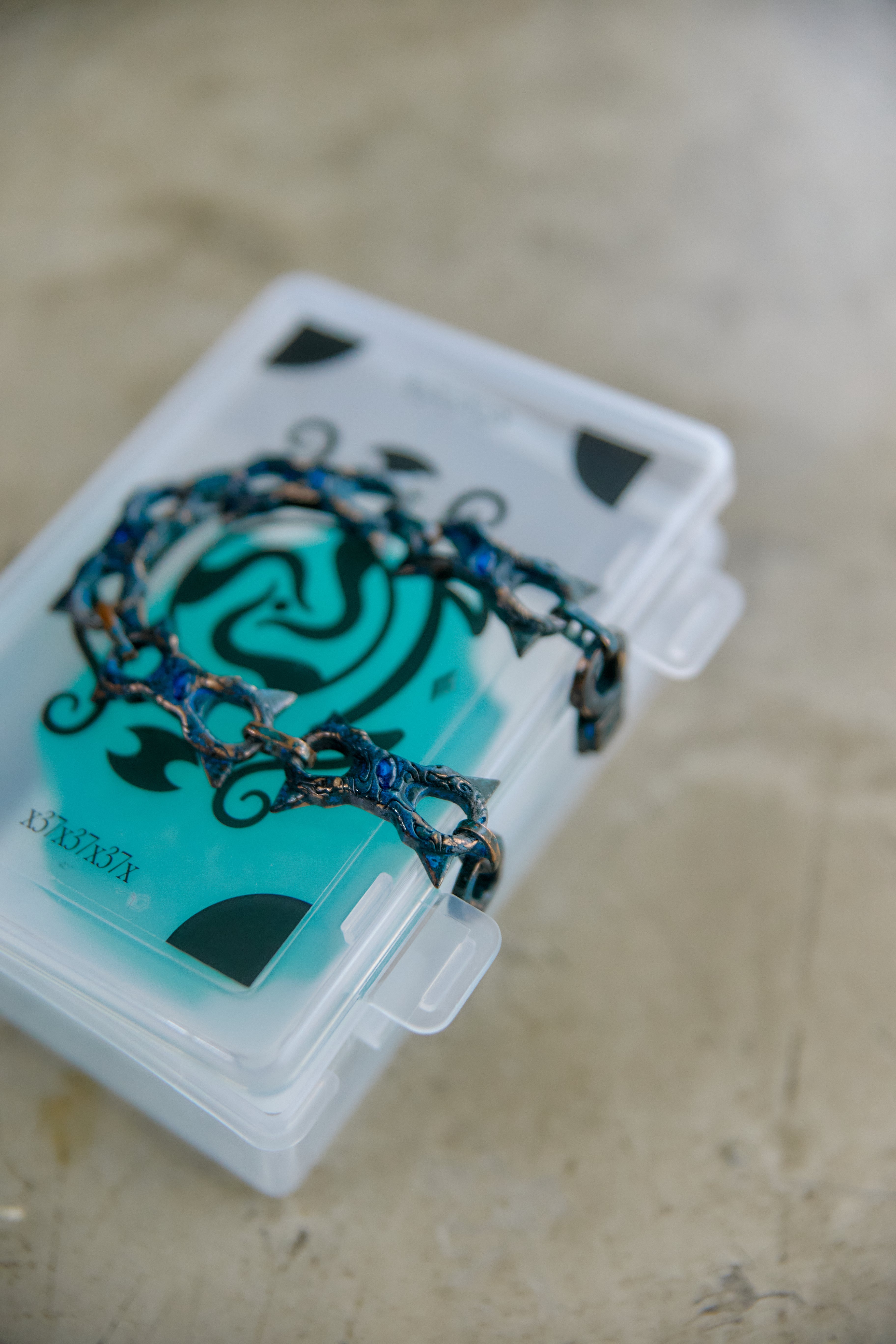 A close-up of a dark metallic chain bracelet with blue and bronze details, resting on a translucent acrylic jewelry box containing a turquoise card, set against a neutral beige background.