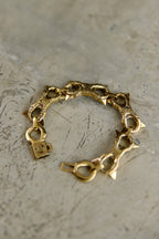 A gold-toned chain bracelet with textured links, triangular spikes, and a small padlock charm, displayed on a textured gray surface.