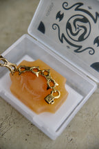 A gold-toned chain accessory with a small padlock charm resting on an orange translucent soap block inside a clear plastic case with a printed black lid design.