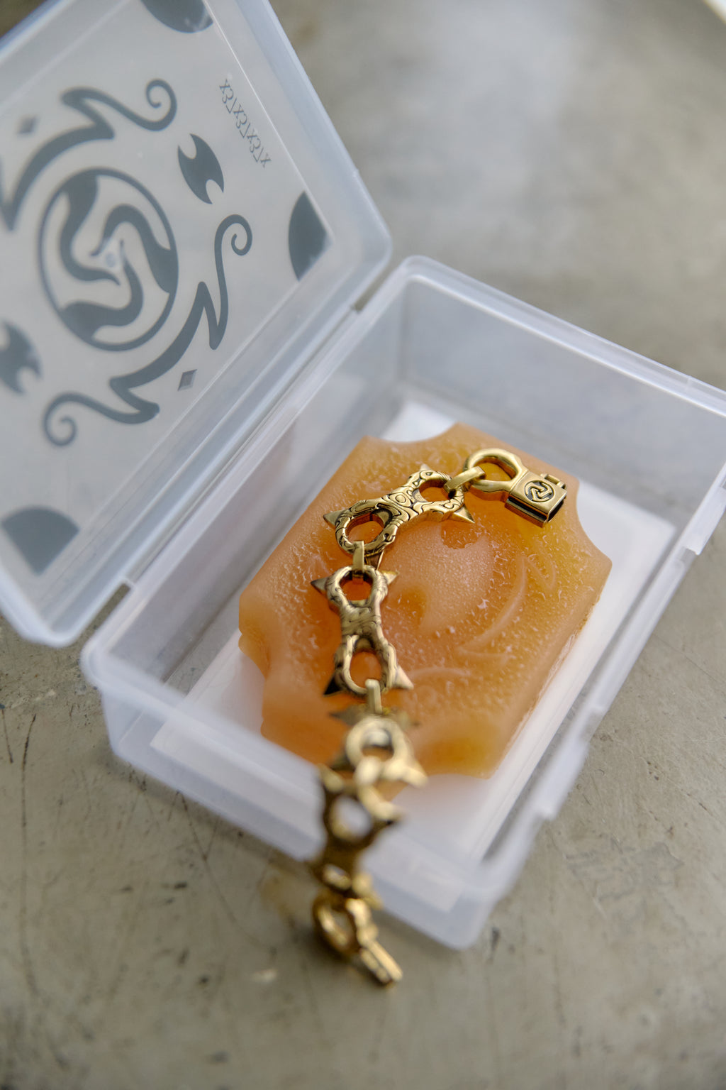 A gold-toned chain bracelet with a small engraved charm, displayed inside a clear plastic storage case with a white insert and decorative lid, set on a gray surface.