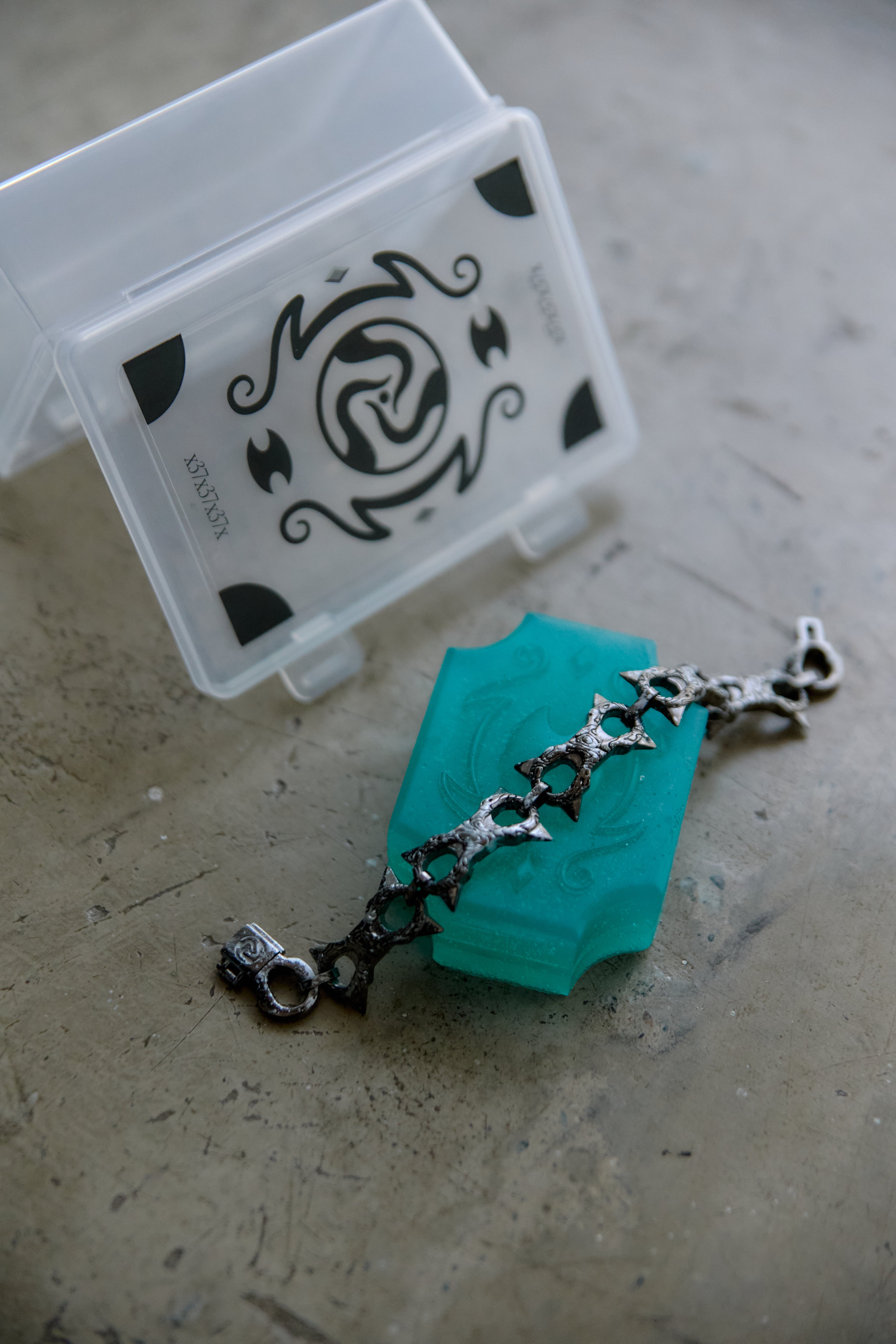 A silver-toned chain bracelet with star-shaped links lies on a textured gray surface next to a translucent turquoise soap block and a clear plastic storage box with a black decorative print.