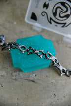 A close-up of a silver-toned metal chain bracelet draped over a translucent turquoise-colored hexagonal soap block, with a blurred white plastic case in the background on a textured gray surface.