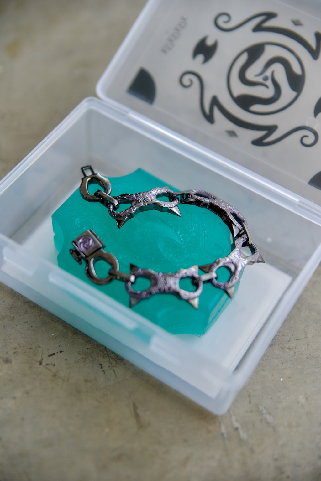 A dark metallic gothic-style chain bracelet with barbed links and a small purple gemstone clasp, resting on a turquoise block inside a clear plastic jewelry box.