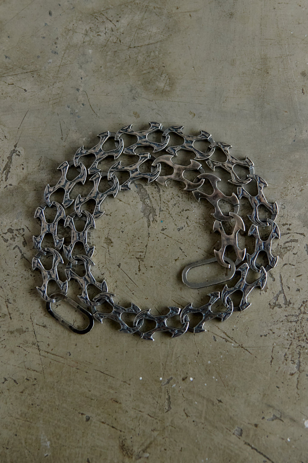 A coiled dark gray metal chain with interlocking links and a small loop at one end, placed on a textured beige concrete-like surface.