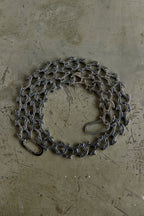 A coiled dark gray metal chain with interlocking links and a small loop at one end, placed on a textured beige concrete-like surface.