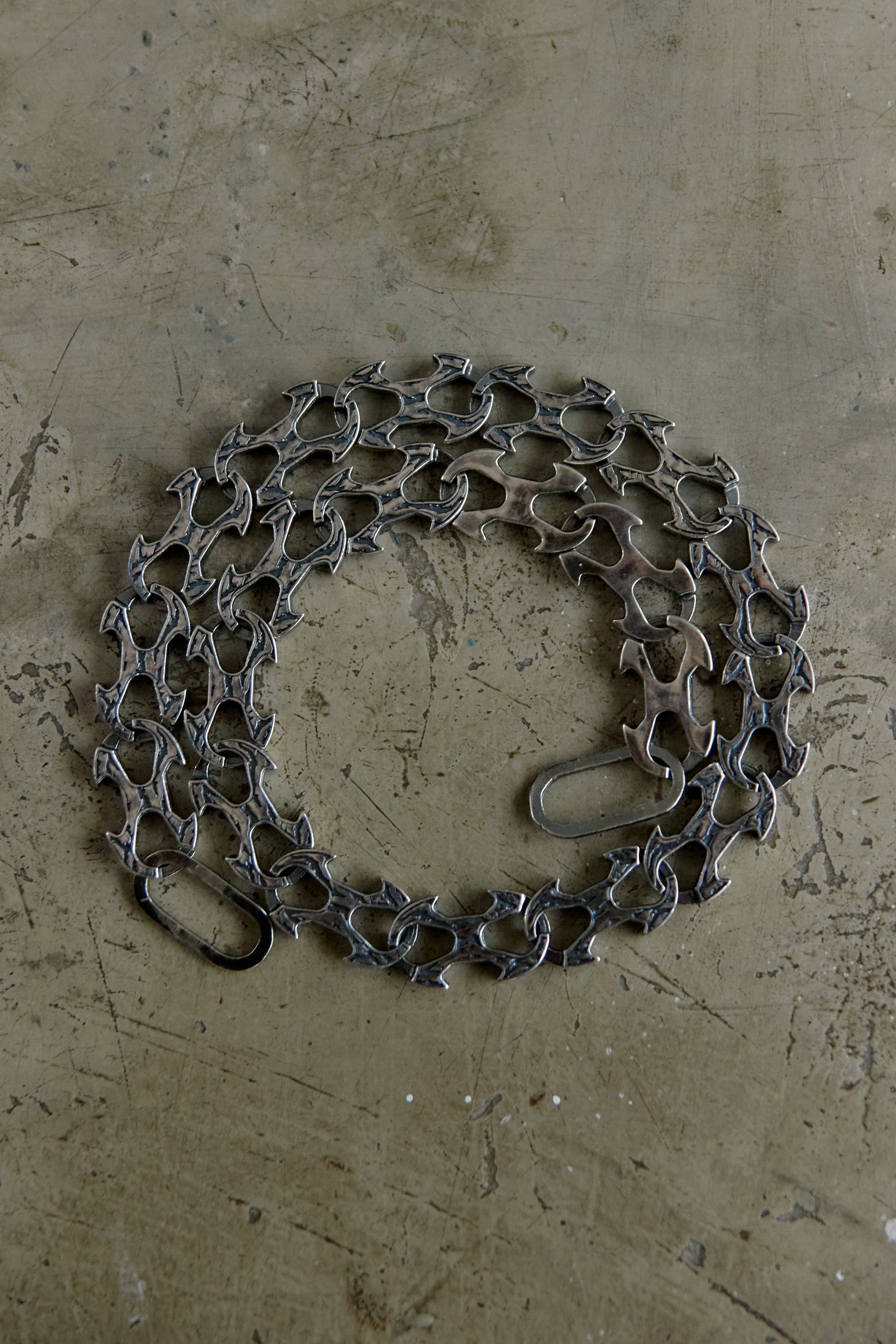 A coiled dark gray metal chain with interlocking links and a small loop at one end, placed on a textured beige concrete-like surface.