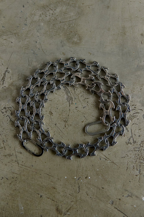 A coiled dark gray metal chain with interlocking links and a small loop at one end, placed on a textured beige concrete-like surface.