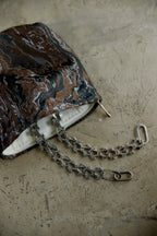A close-up of a handbag with a marbled black, brown, and copper patterned exterior, featuring a silver-tone chain strap and a white interior lining, resting on a textured concrete surface.