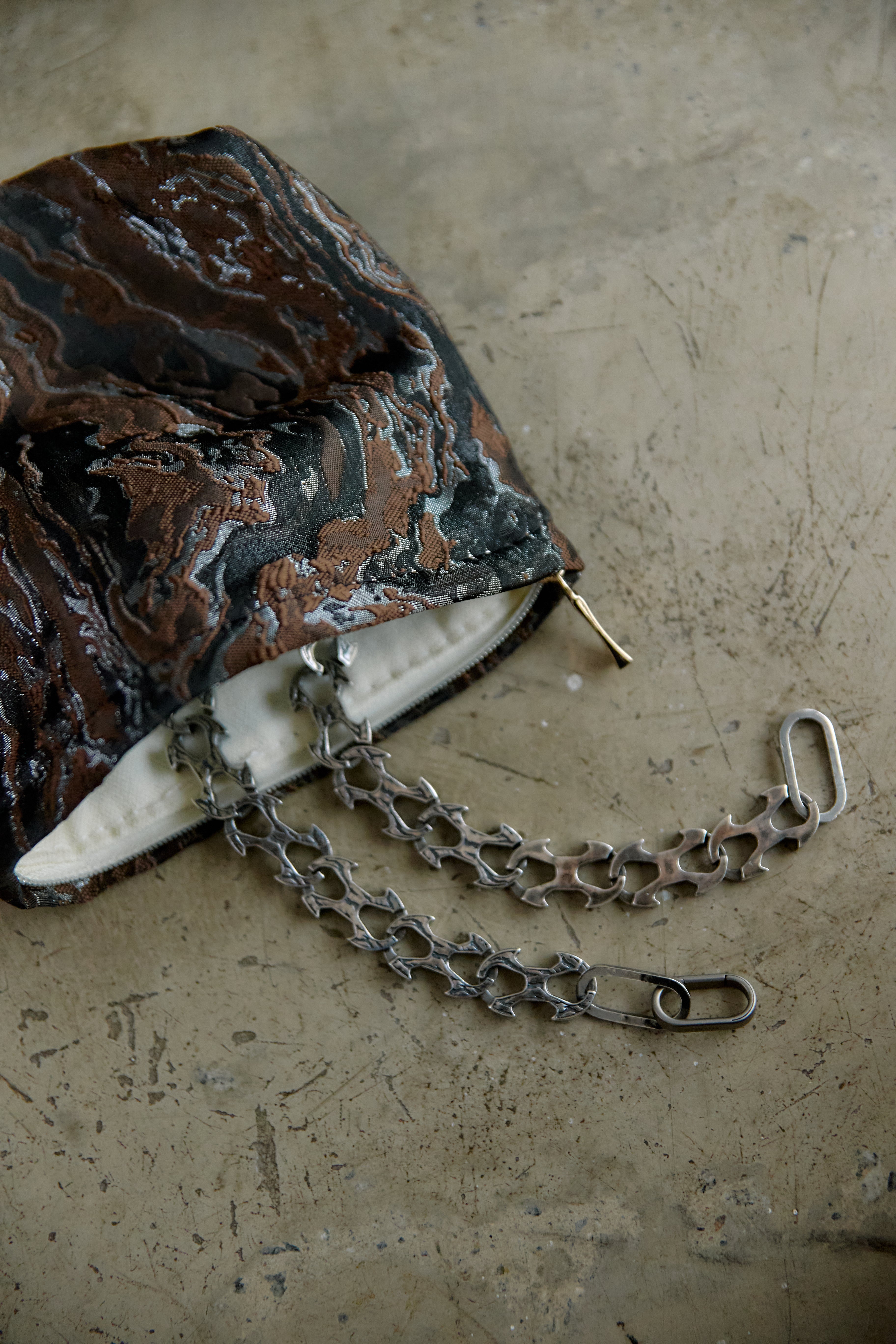 A close-up of a handbag with a marbled black, brown, and copper patterned exterior, featuring a silver-tone chain strap and a white interior lining, resting on a textured concrete surface.