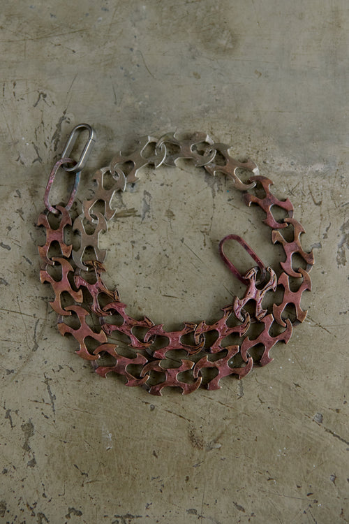A bicycle chain guard with a reddish-brown protective plate and silver metal chain links, laid flat on a textured concrete surface.