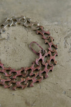 A close-up of a vintage-style metal chain necklace with interlocking cutout links, featuring a reddish-pink patina and silver-toned links, resting on a textured concrete surface.