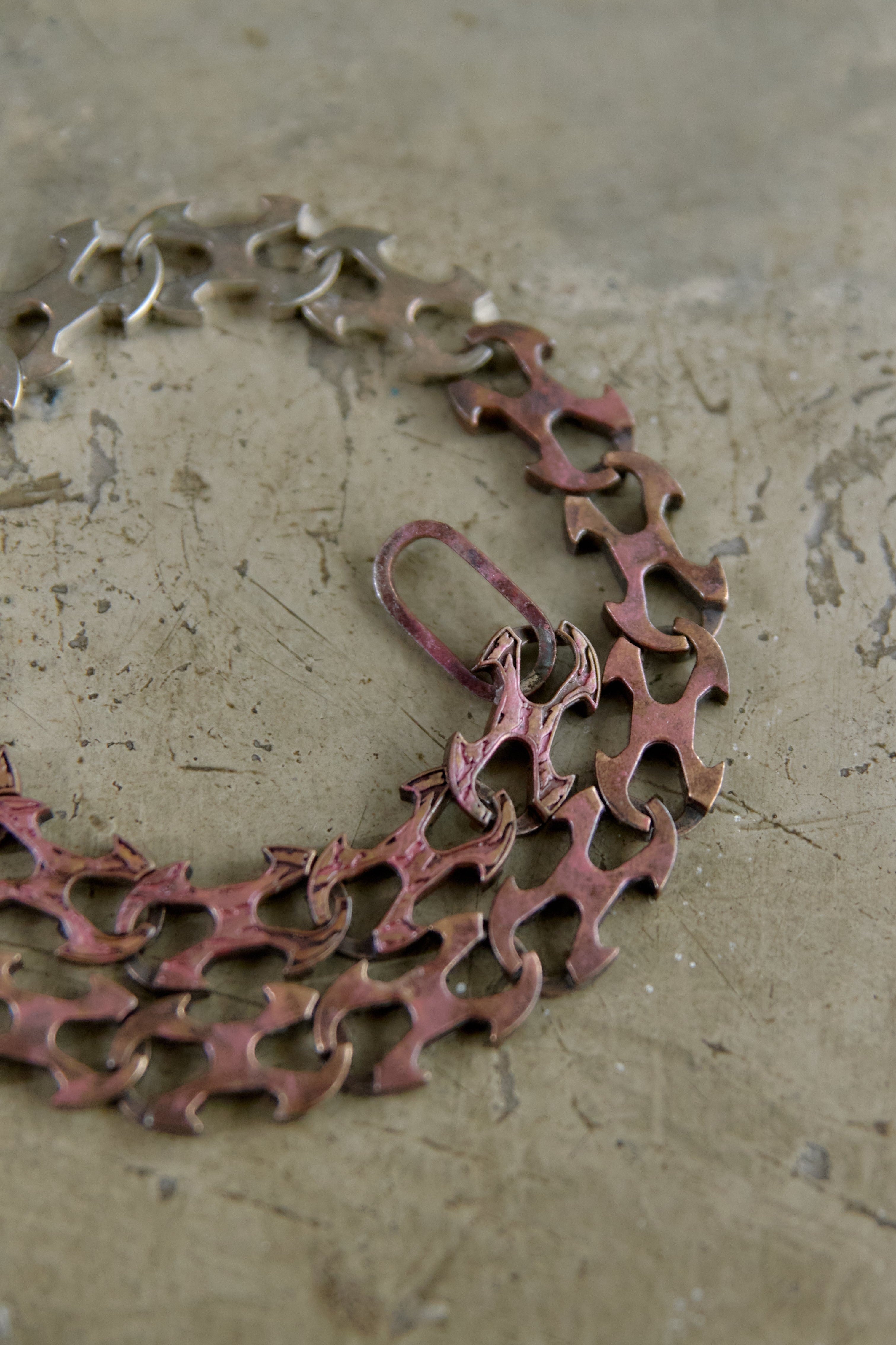 A close-up of a vintage-style metal chain necklace with interlocking cutout links, featuring a reddish-pink patina and silver-toned links, resting on a textured concrete surface.