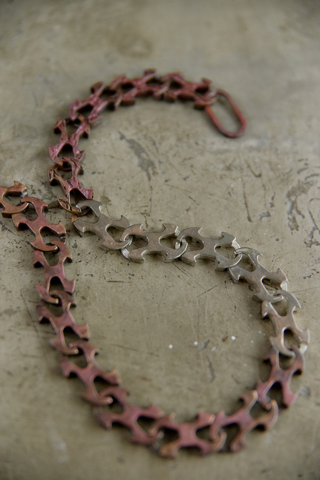 A handmade link chain necklace made from cut bicycle chainstays, featuring a rusted reddish-brown finish on one side and a silver-toned finish on the other, displayed on a textured concrete surface.