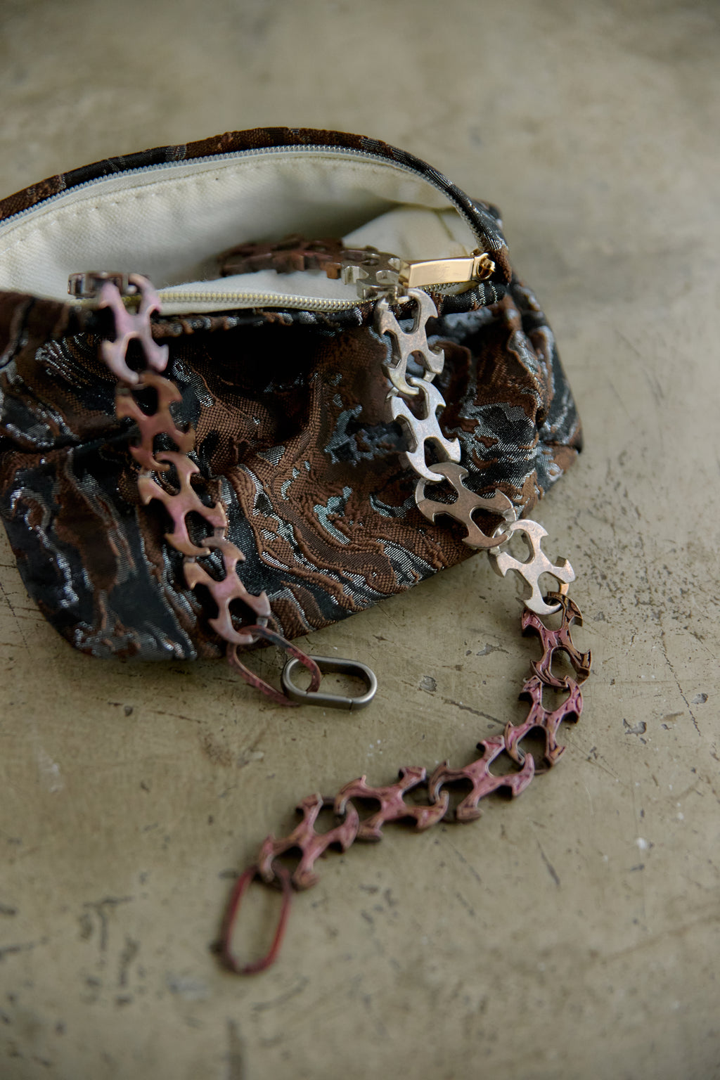 A close-up of a small zippered pouch with a marbled brown, black, and gray pattern, featuring a decorative chain strap made of bicycle chainrings in pink, silver, and rose gold tones, resting on a textured concrete surface.
