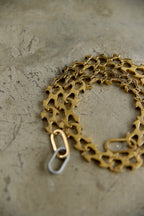A close-up of a gold-tone bicycle chain-link style bracelet or necklace laid on a textured gray surface, with a small silver-toned clasp visible.