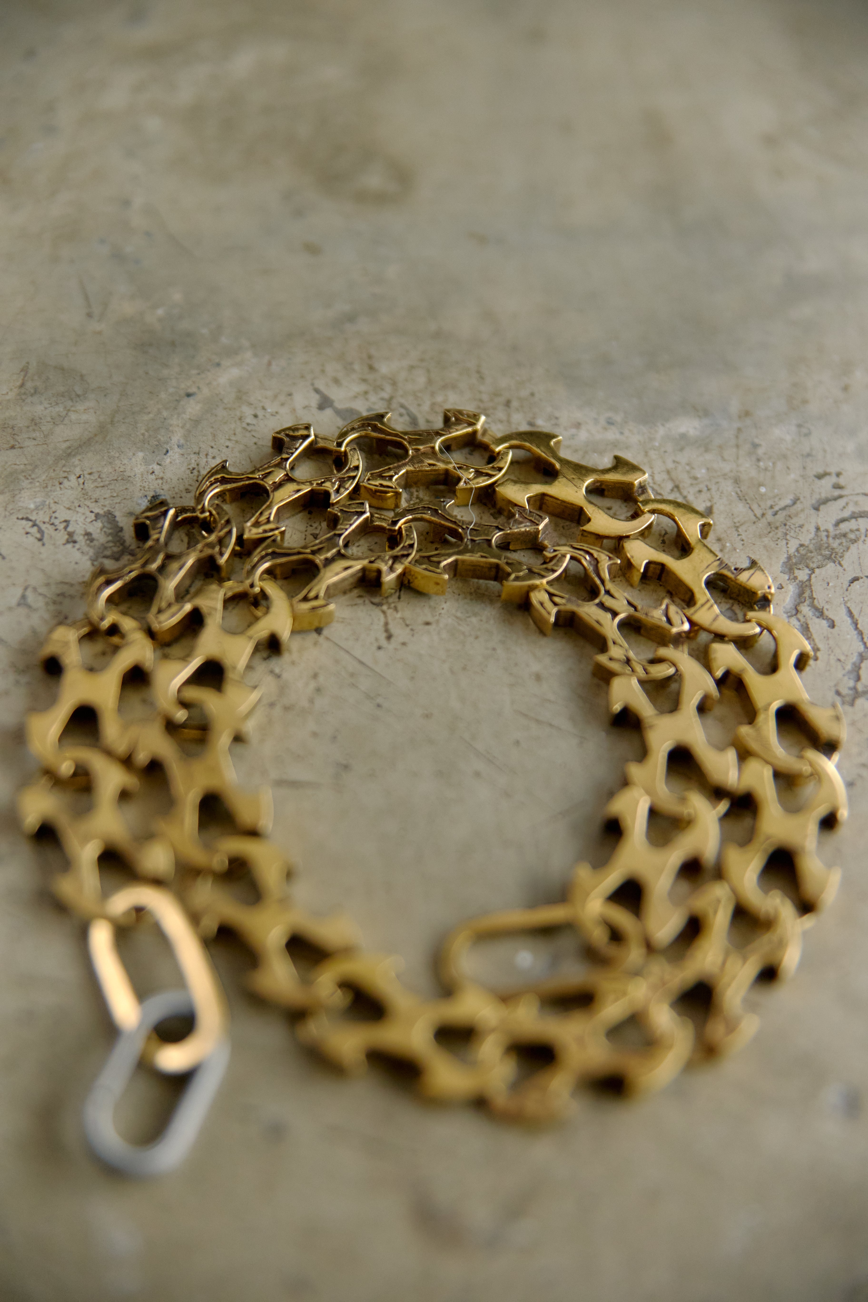 A gold-toned metal chain necklace with a unique openwork, gear-like link pattern, coiled on a textured gray concrete surface.
