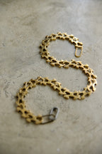 A gold-toned chain bracelet with a unique openwork, gear-like link design, displayed on a textured concrete surface.