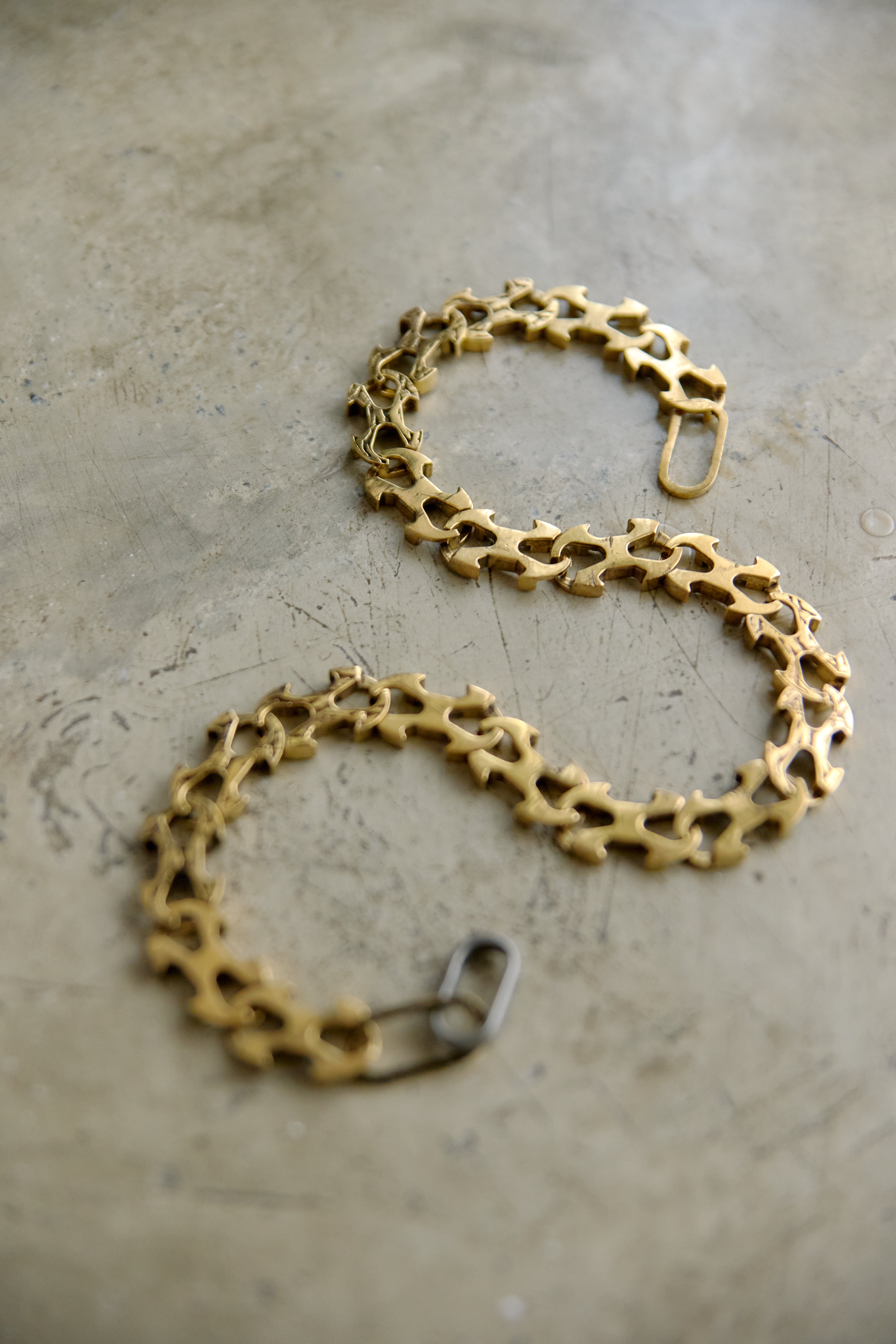 A gold-toned chain bracelet with a unique openwork, gear-like link design, displayed on a textured concrete surface.