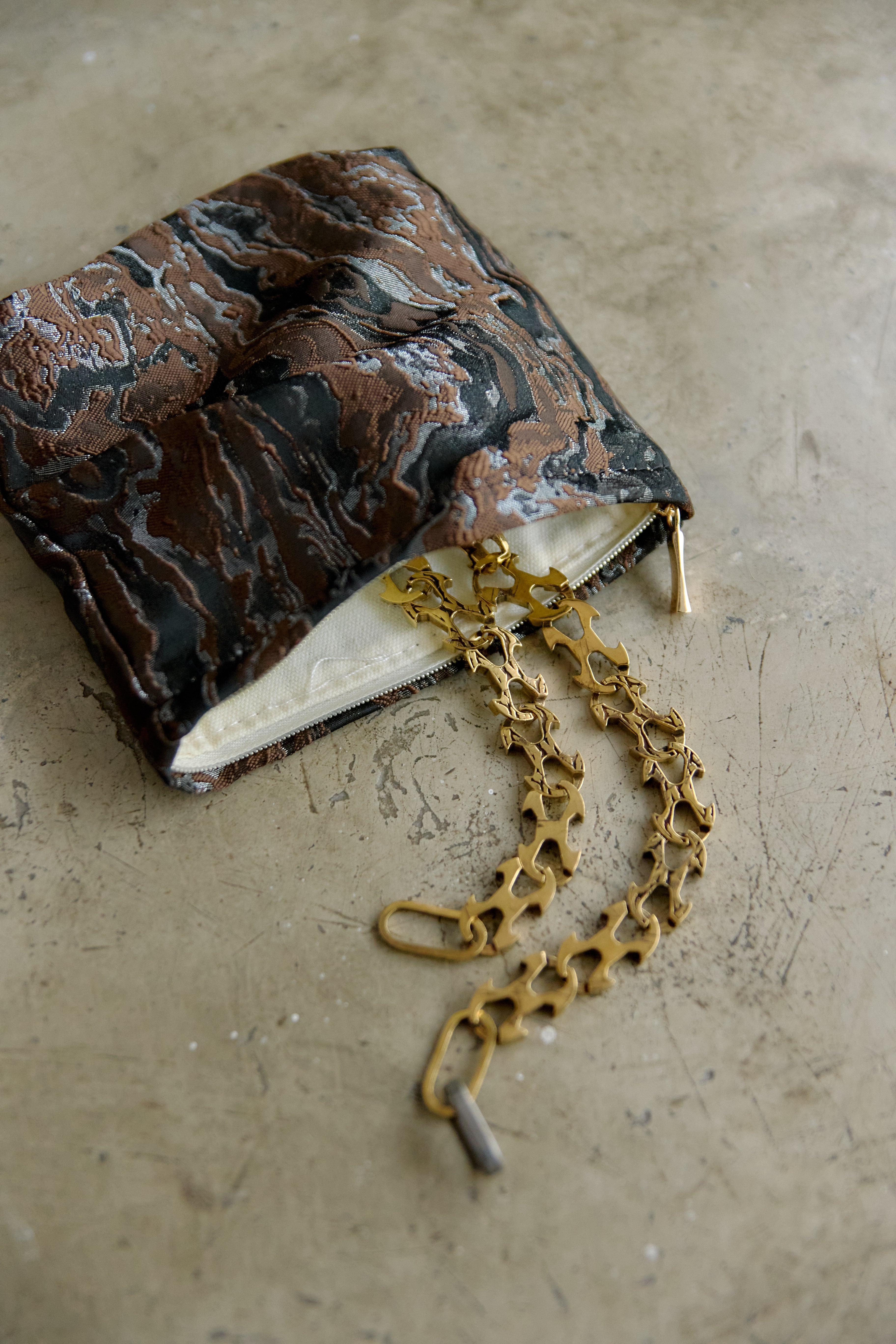 A small marbled brown and black fabric pouch with a gold-tone chain and clasp, photographed on a textured beige surface.