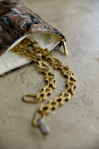 A close-up of a gold chain necklace draped from an open patterned pouch with a cream lining, placed on a textured gray surface.