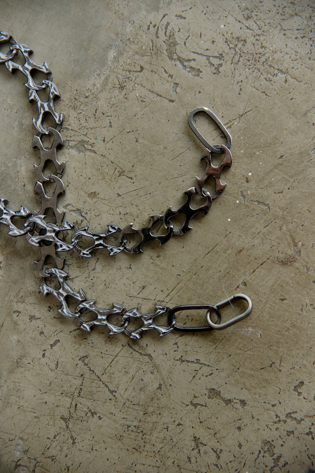A handmade metal chain accessory with a unique textured, hammered finish, laid out on a textured concrete surface.