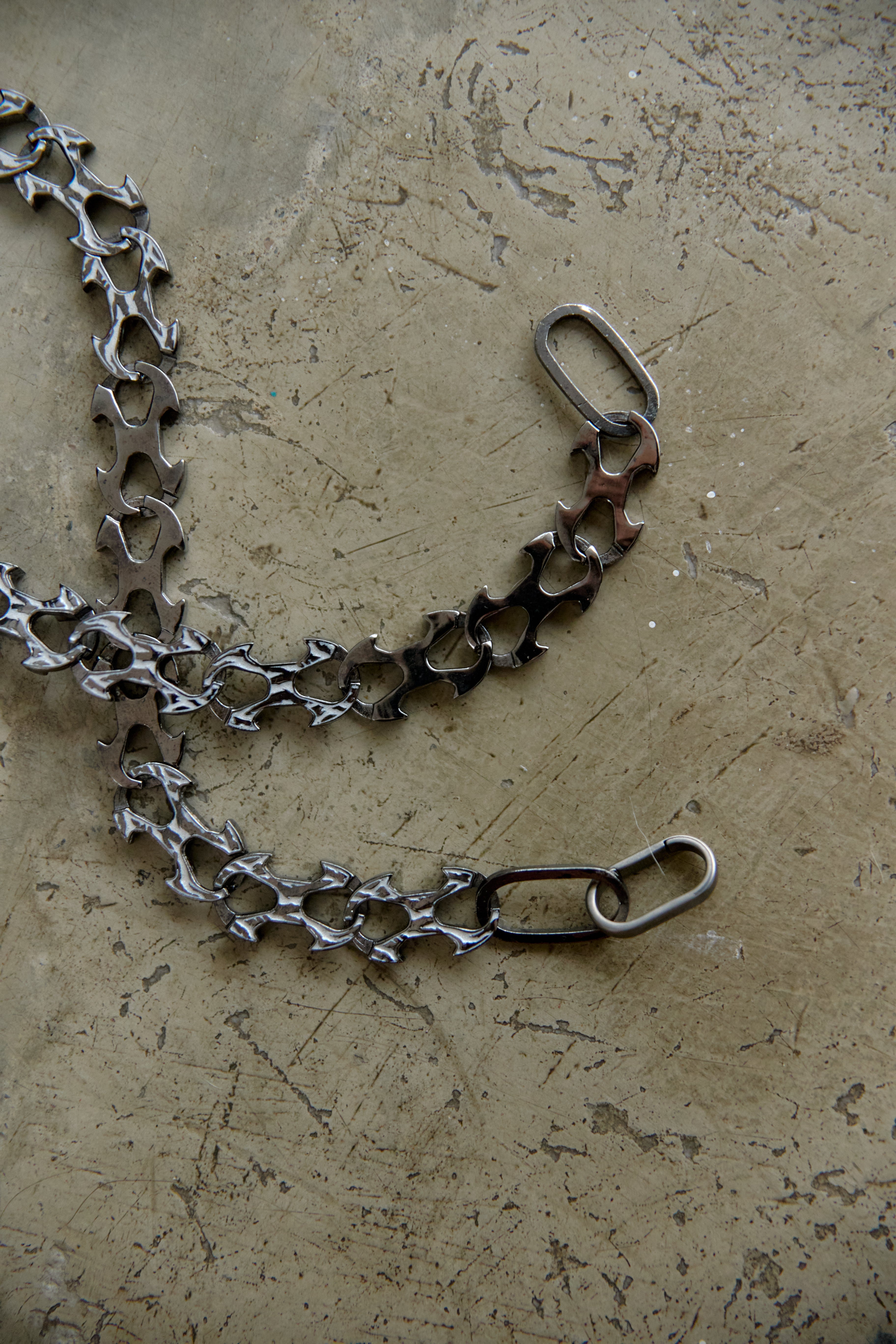 A handmade metal chain accessory with a unique textured, hammered finish, laid out on a textured concrete surface.
