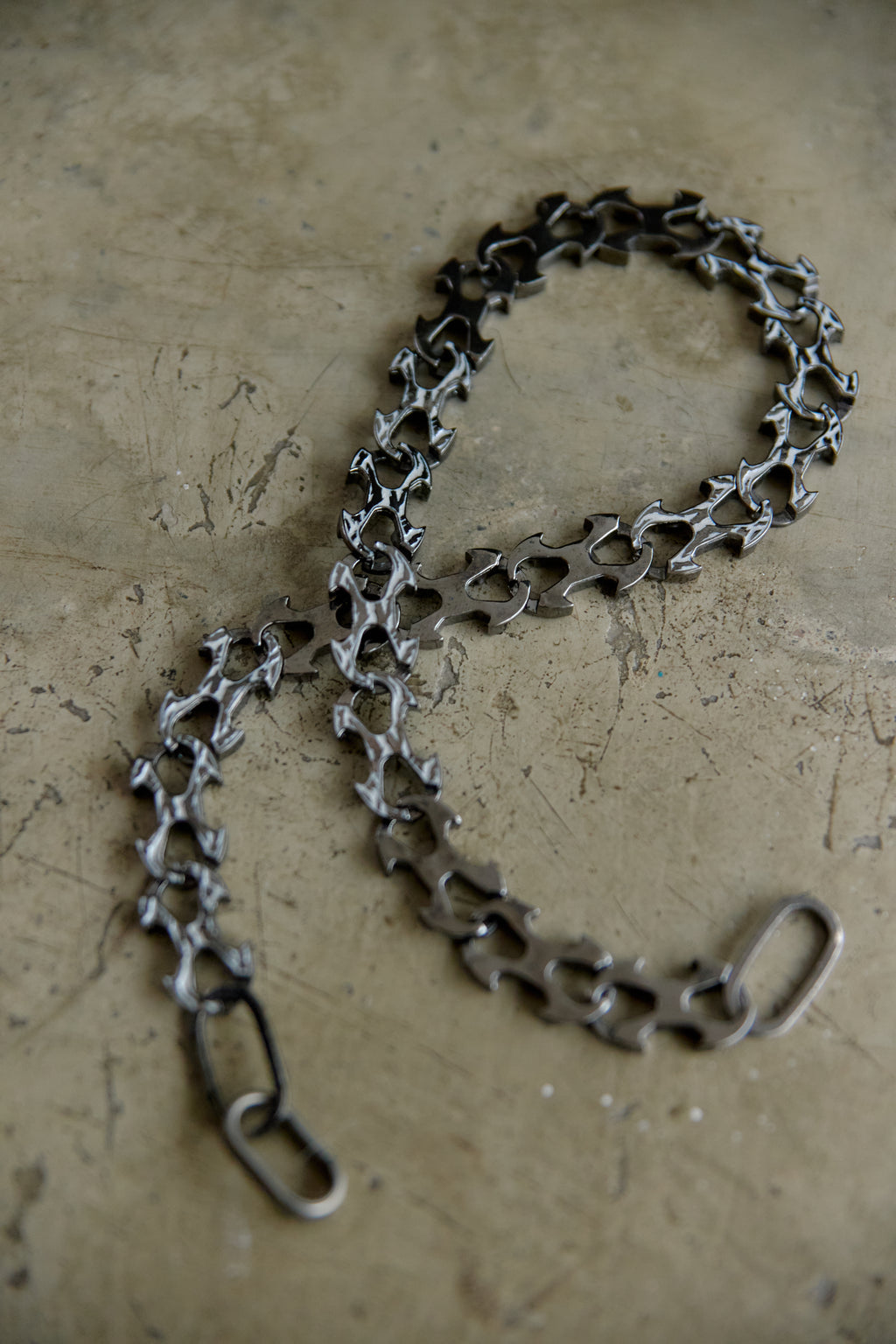 A dark metallic bicycle chain link chain with a matching rear hook, laid out on a textured concrete surface.