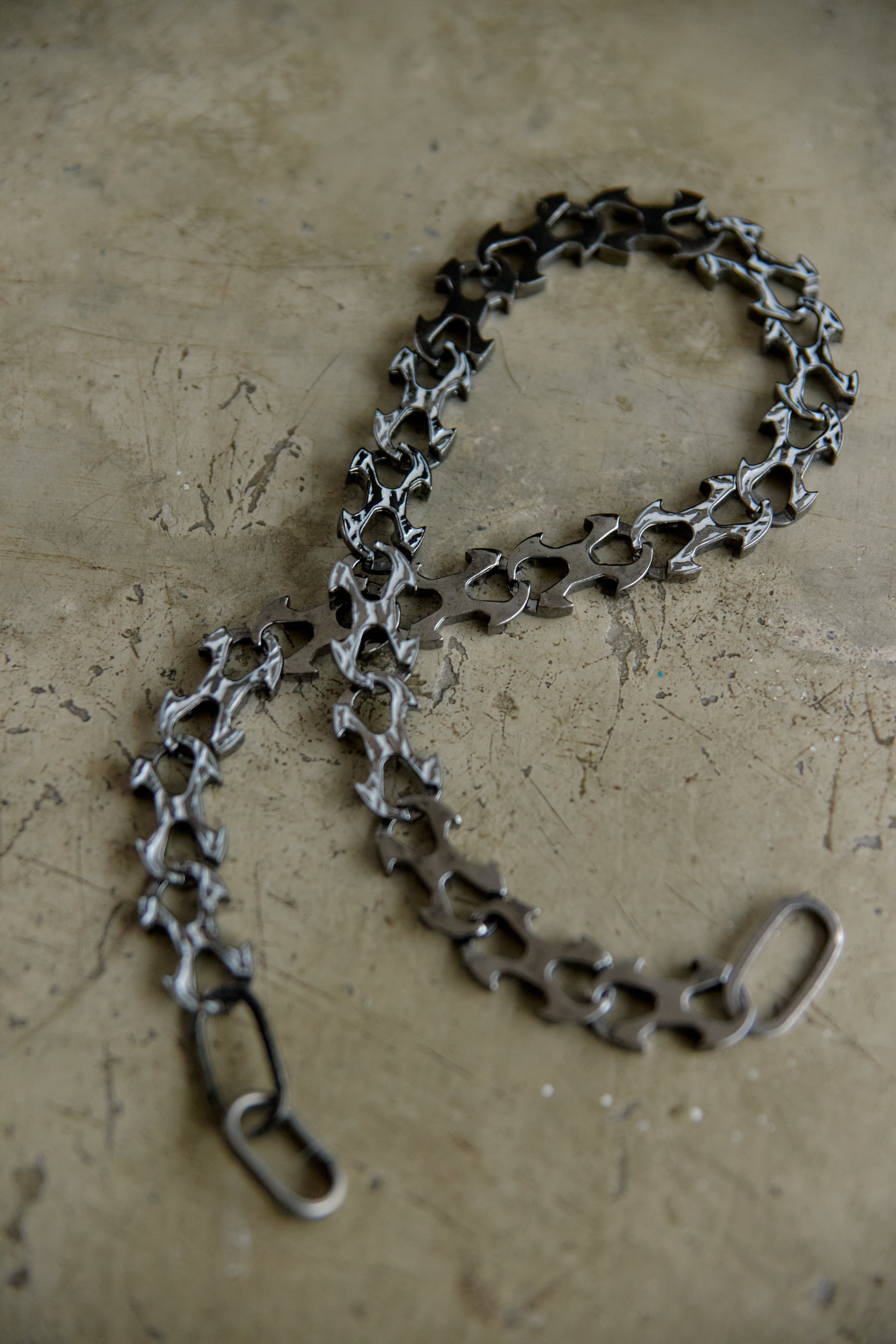 A dark metallic bicycle chain link chain with a matching rear hook, laid out on a textured concrete surface.