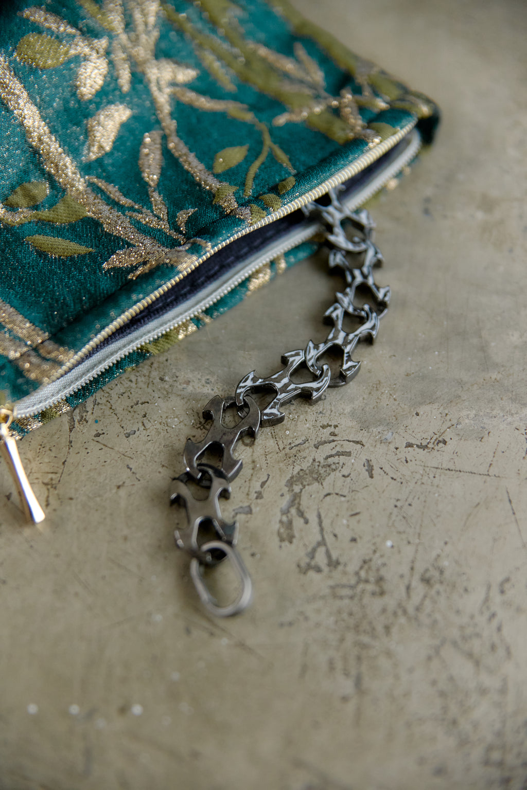 A close-up of a teal and gold patterned fabric zip pouch with a dark metallic ornate key-shaped zipper pull resting on a textured beige surface.
