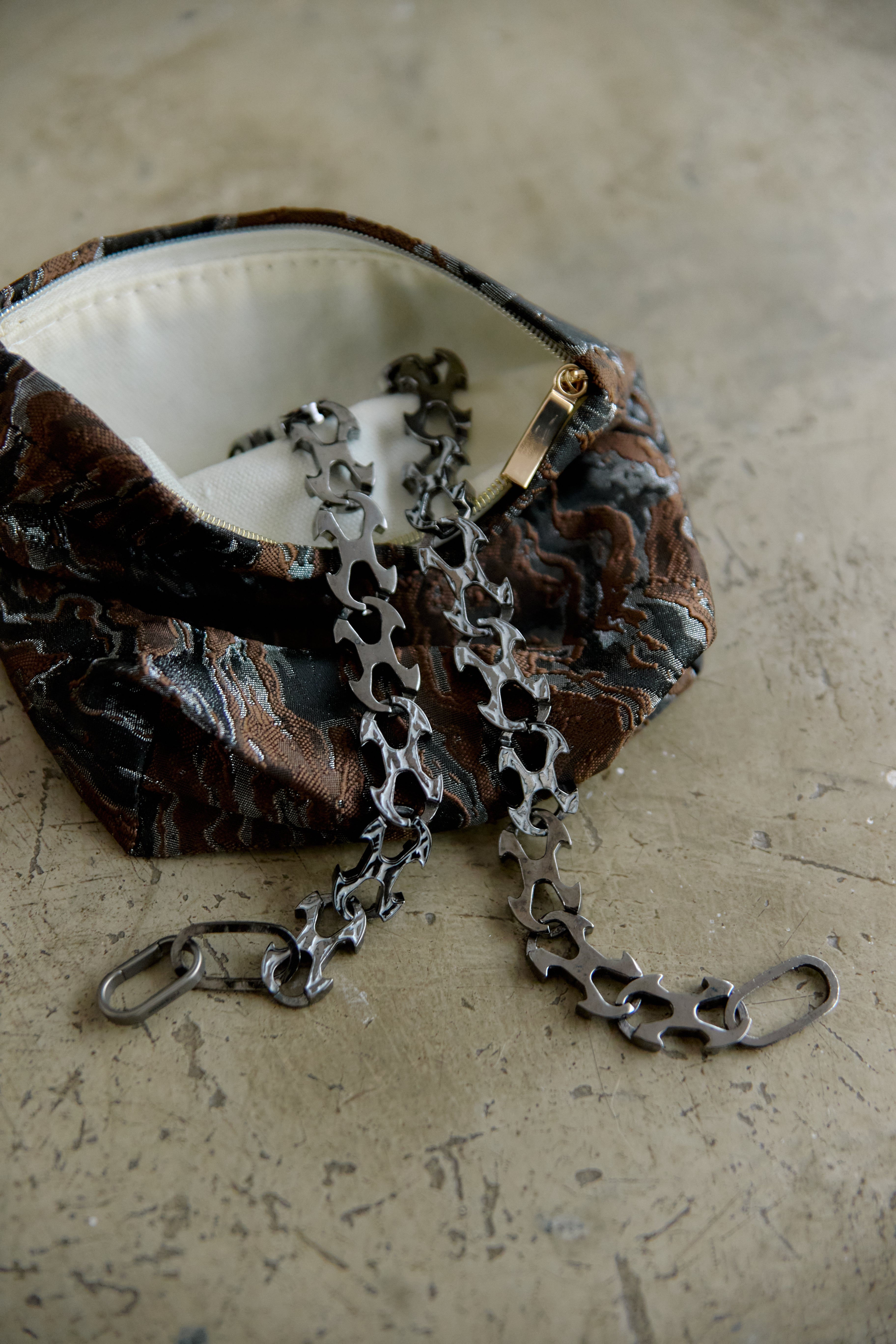 A close-up of a small zippered pouch with a dark brown and black abstract marbled pattern, featuring a silver bicycle chain strap and a gold-tone zipper pull, resting on a textured beige surface.